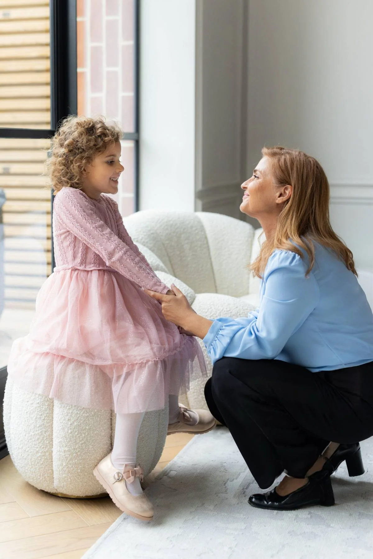A young girl in a pink dress sitting on a round white chair, smiling and holding hands with Stella in a blue blouse who is kneeling in front of her, both looking happy in a bright room with a window.