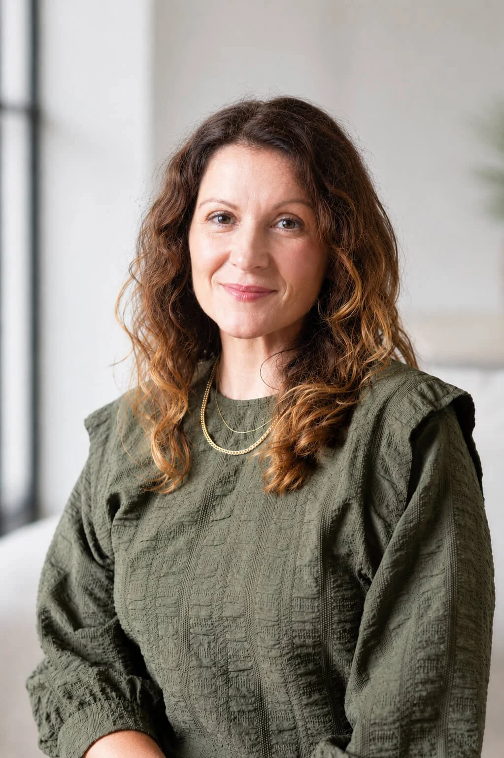 Lana Novak, a woman with curly brown hair and light skin, smiling, wearing an olive green textured blouse and gold jewellery, sitting indoors with natural light coming through large windows.