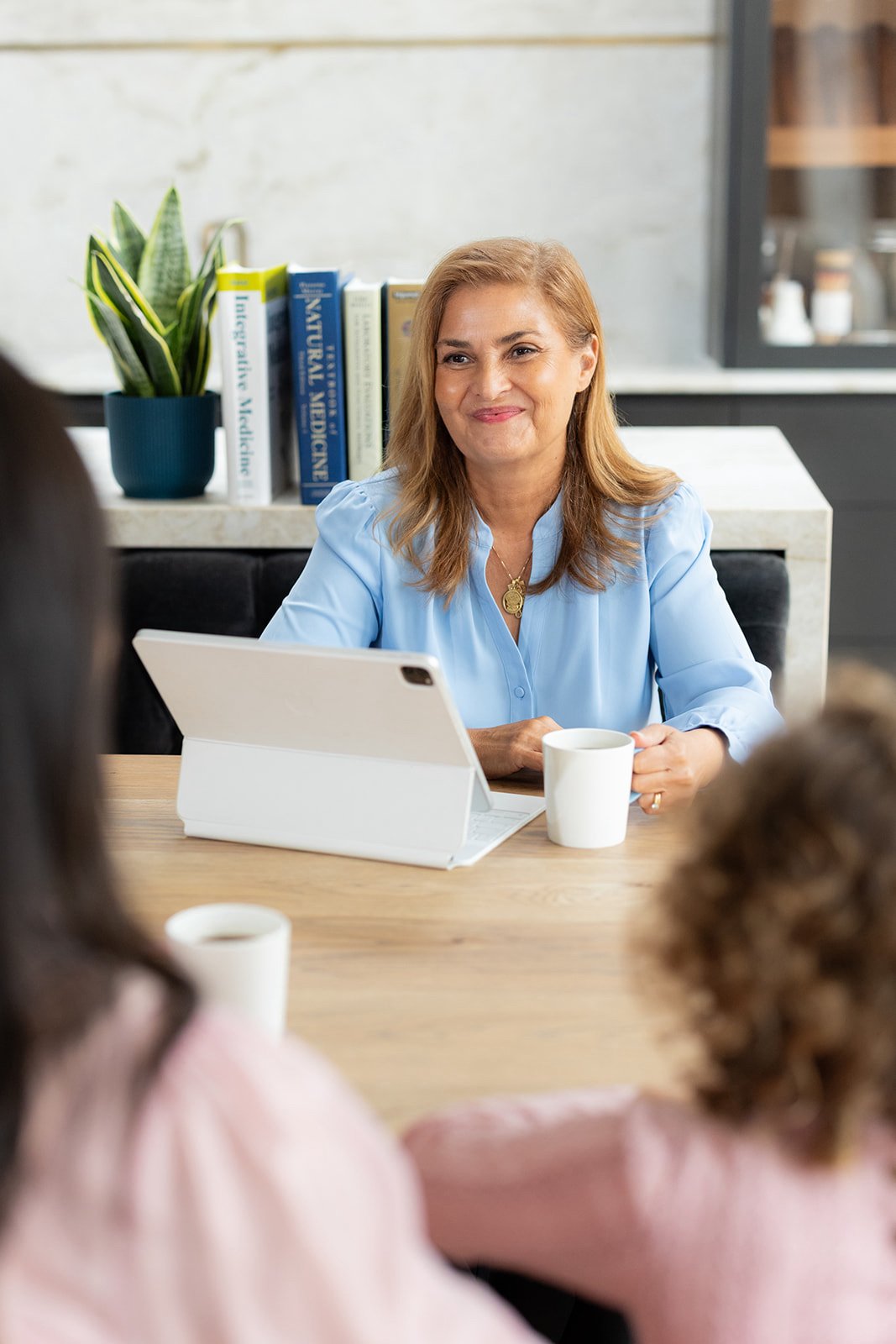 Stella in a blue shirt sitting at a table with a tablet and coffee mug, during a meeting with a mum and her daughter in the foreground.
