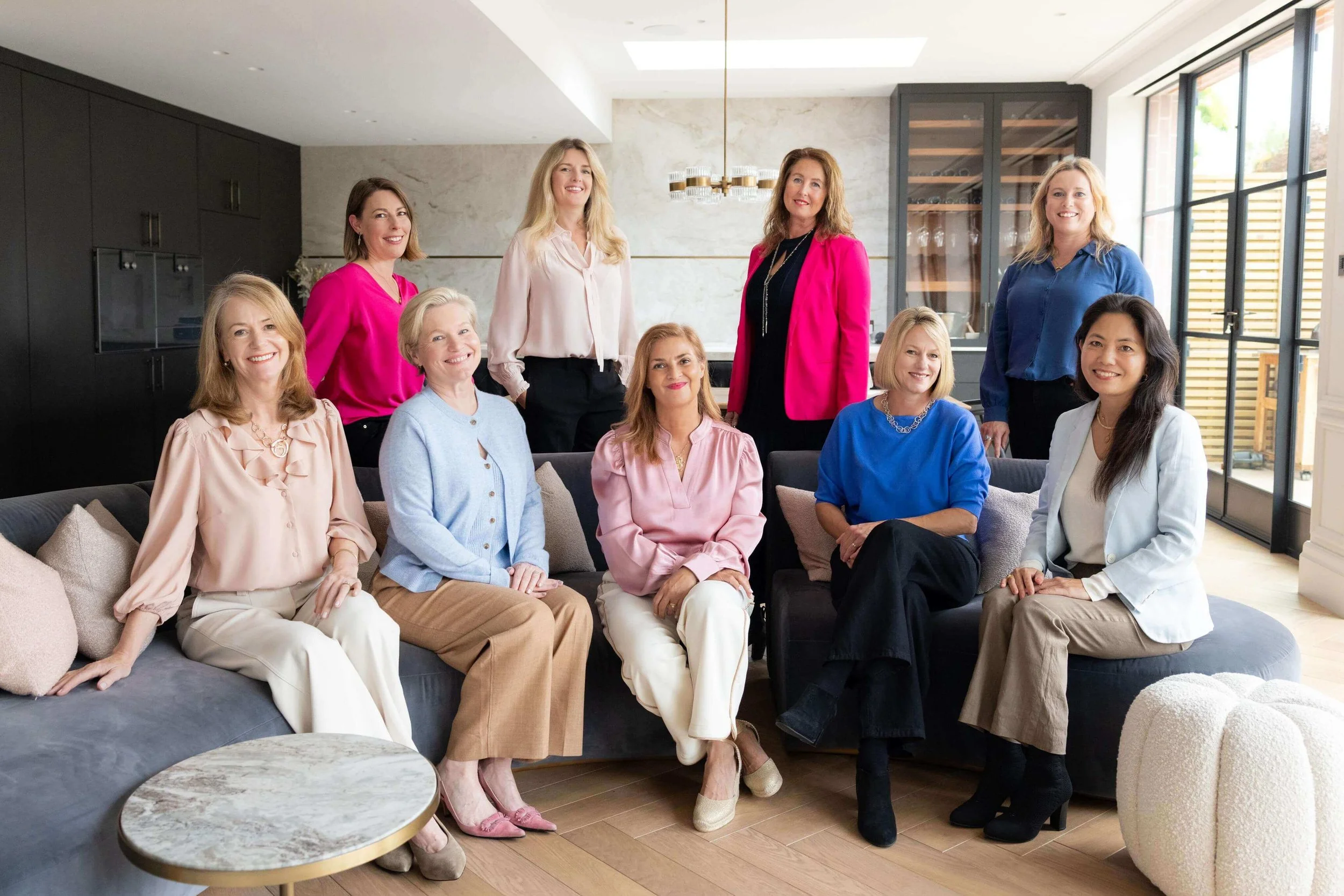 The Brainstorm Health team. Nine women gathered in a modern, well-lit living room, smiling at the camera, with some sitting on a sofa and others standing behind.