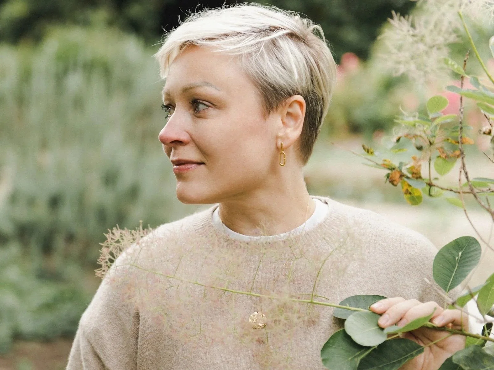 A woman with short blonde hair and light skin wears a light-colored sweater and gold earrings, standing outdoors surrounded by greenery.