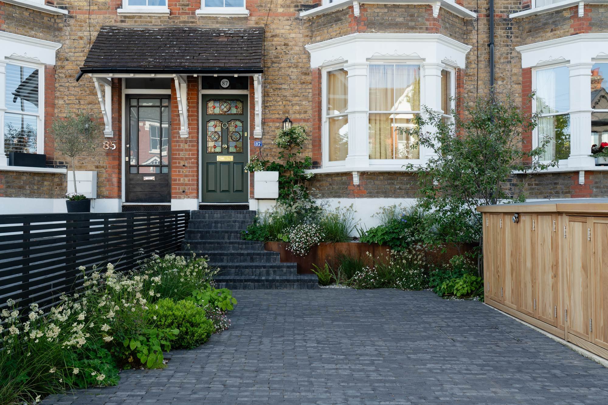 Front of a brick house with a black gate and a small garden in the front yard, featuring a variety of plants and flowers, a black fence on the left, and a wooden storage shed on the right.