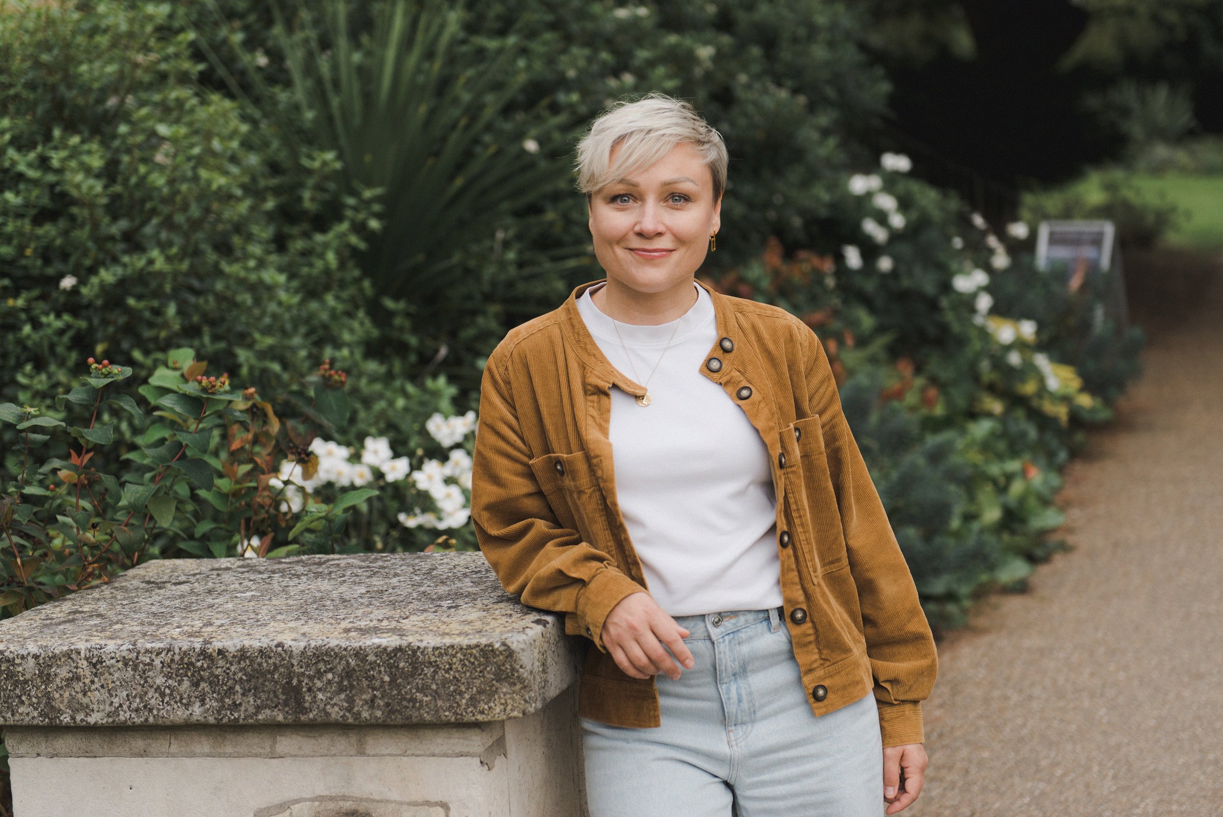 A woman with short blonde hair, wearing a white t-shirt and a brown corduroy jacket, stands next to a stone pillar in a garden with green bushes and white flowers.