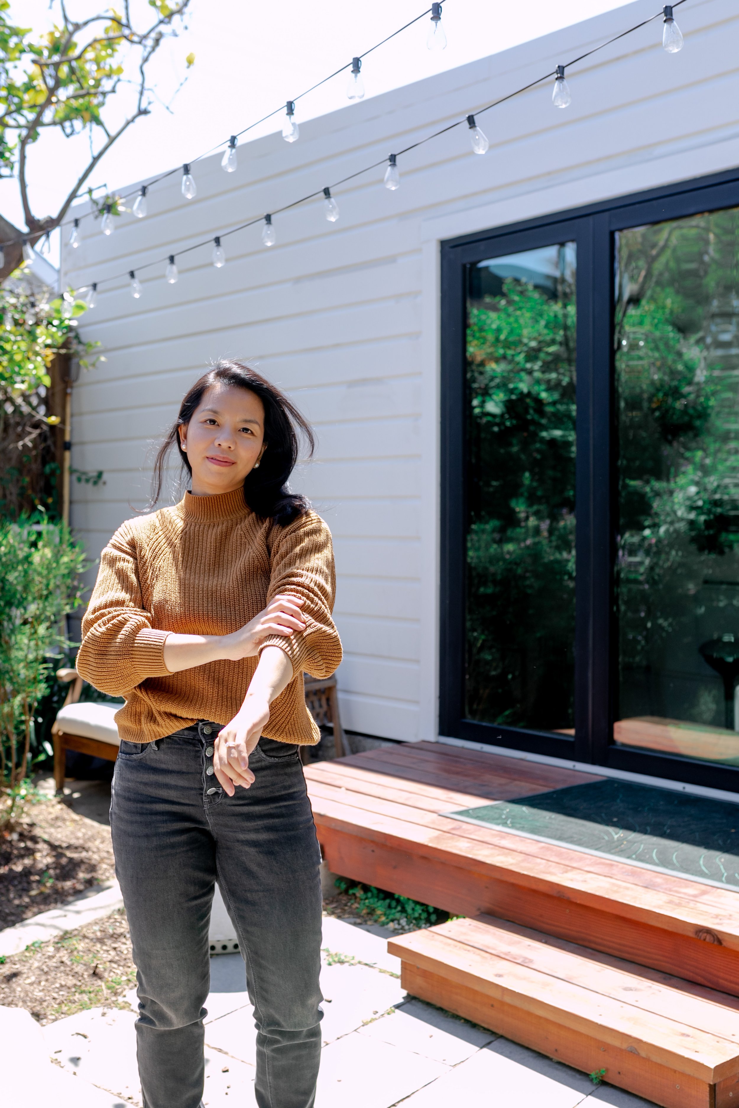A woman standing outside on a sunny day near a white house with black framed doors, string lights overhead, and green foliage.