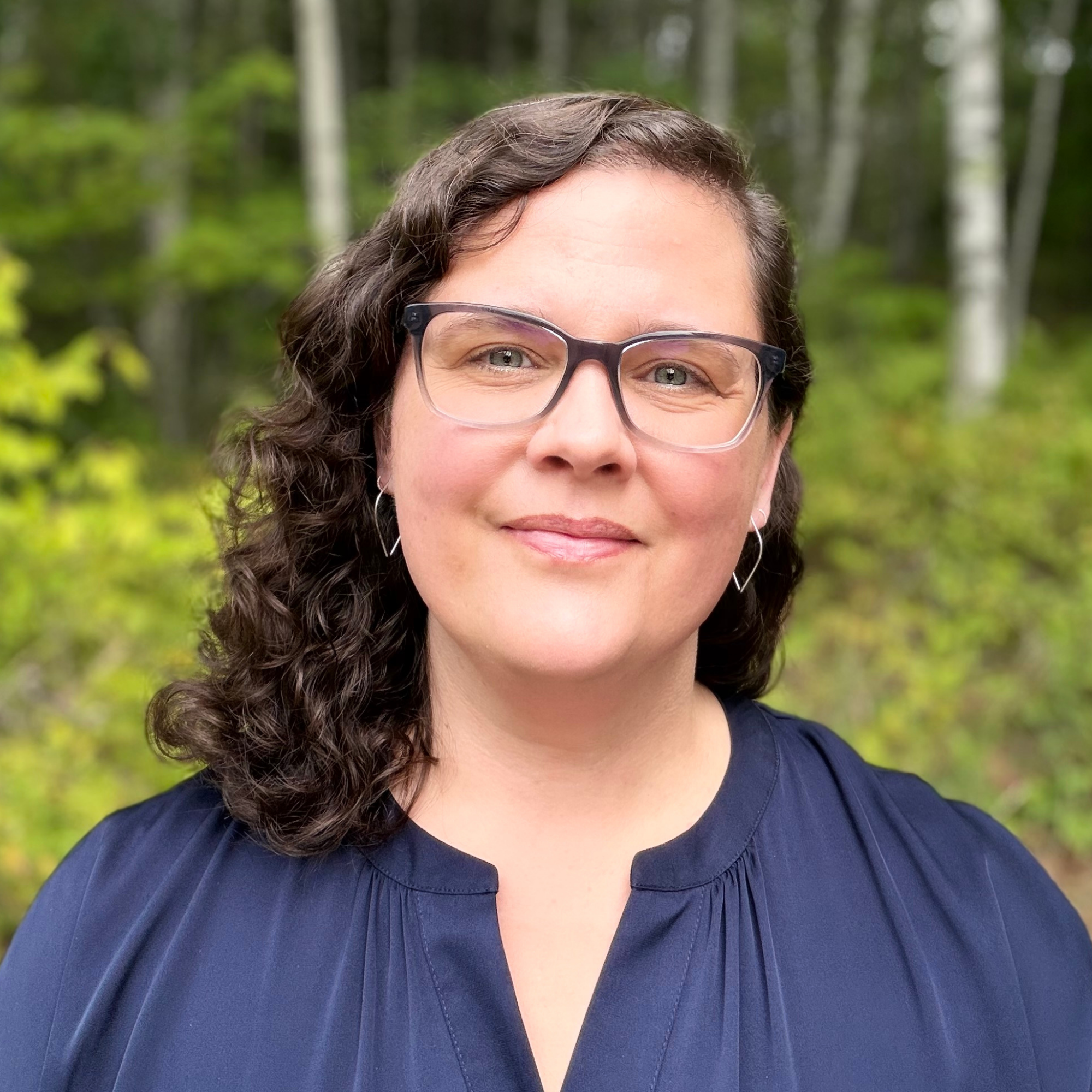 A woman with curly brown hair and glasses standing outdoors in a garden with trees and green grass.