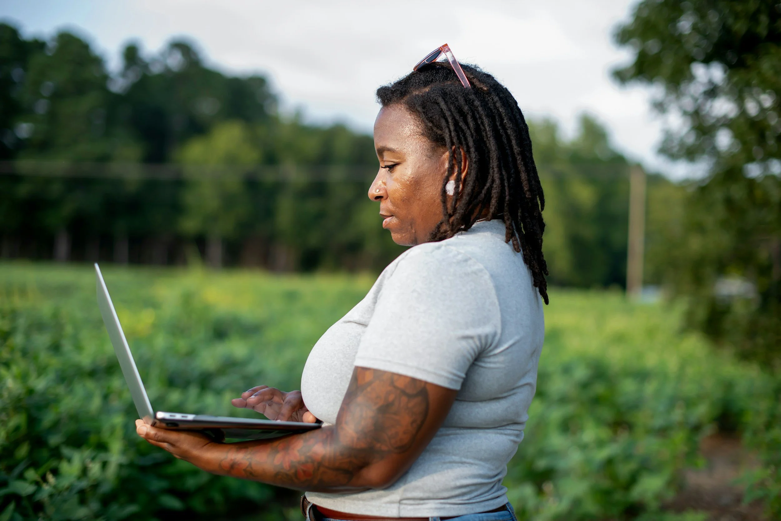 A woman with dreadlocks and tattoos is outdoors, looking at a laptop she is holding in her hands, with a background of green trees and open sky.