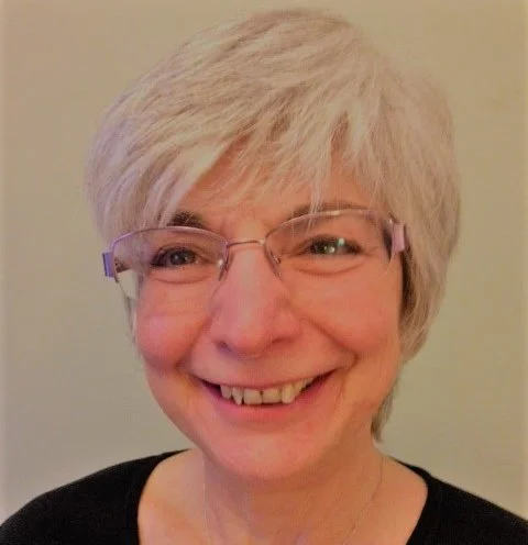 Close-up of a smiling woman with short white hair and purple glasses, wearing a black top.