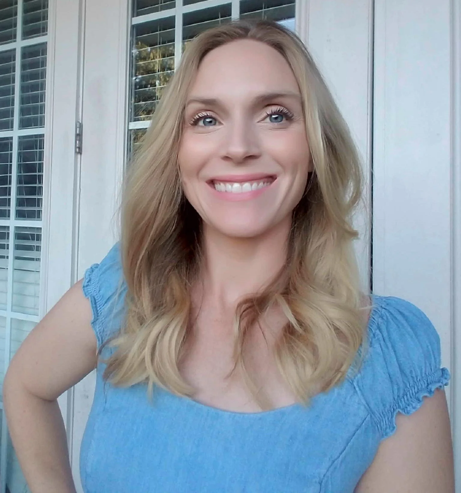 Close-up of smiling woman with long blonde hair, wearing a blue top, standing outside in front of a building with white shutters and large glass windows.