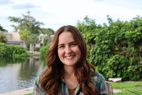 A young woman with long brown hair smiling outdoors near a body of water and green trees.