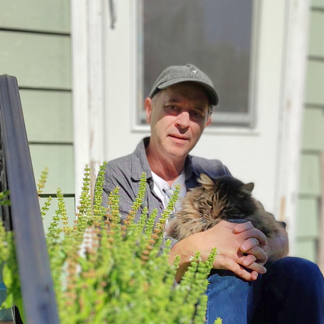 Man seated in the sun with a baseball hat, holding a cat in his lap