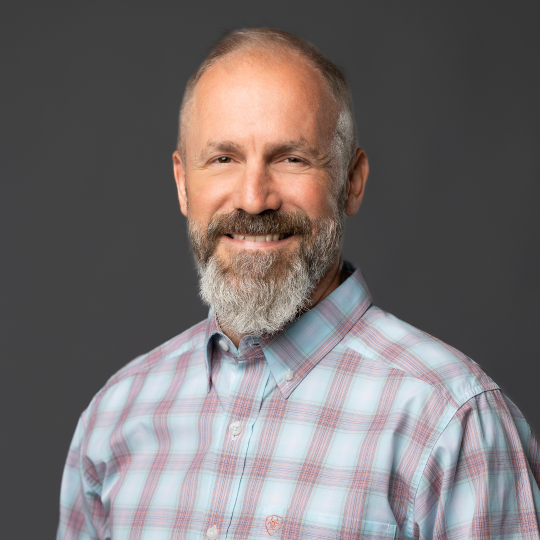 Headshot of a smiling middle-aged man with a beard and mustache, wearing a plaid button-up shirt, against a solid gray background.