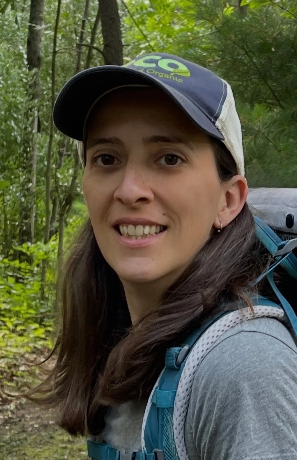 A young woman with long brown hair wearing a camouflage cap, sunglasses, a black and white plaid shirt, and blue jeans, sitting near a river with trees in autumn colors in the background.