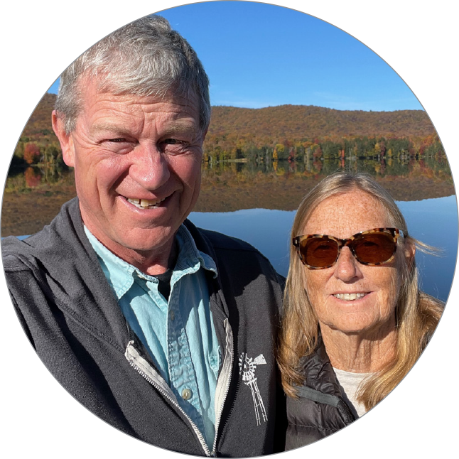 A smiling man and woman taking a selfie outdoors by a lake with colorful autumn trees and a hill in the background.