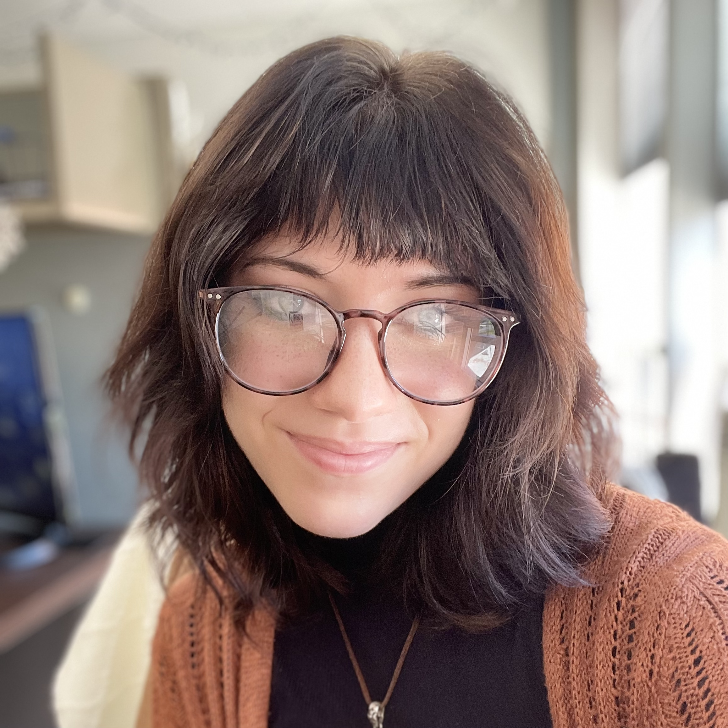 A woman with dark hair, glasses, and a septum piercing smiling at the camera, sitting in a warmly lit room.