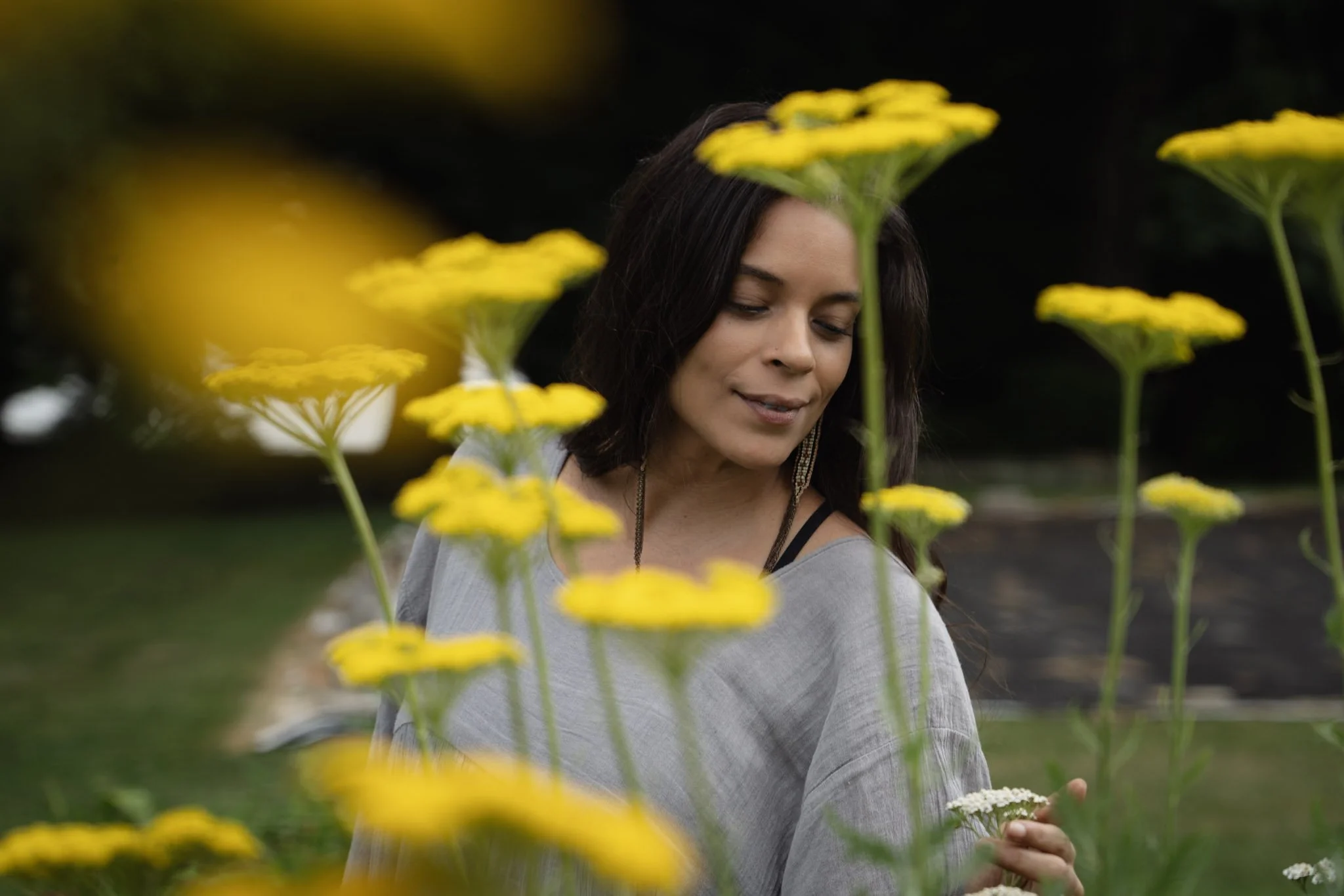 A woman with dark hair and a gray top standing among yellow flowers in a garden or park, with a dark background.