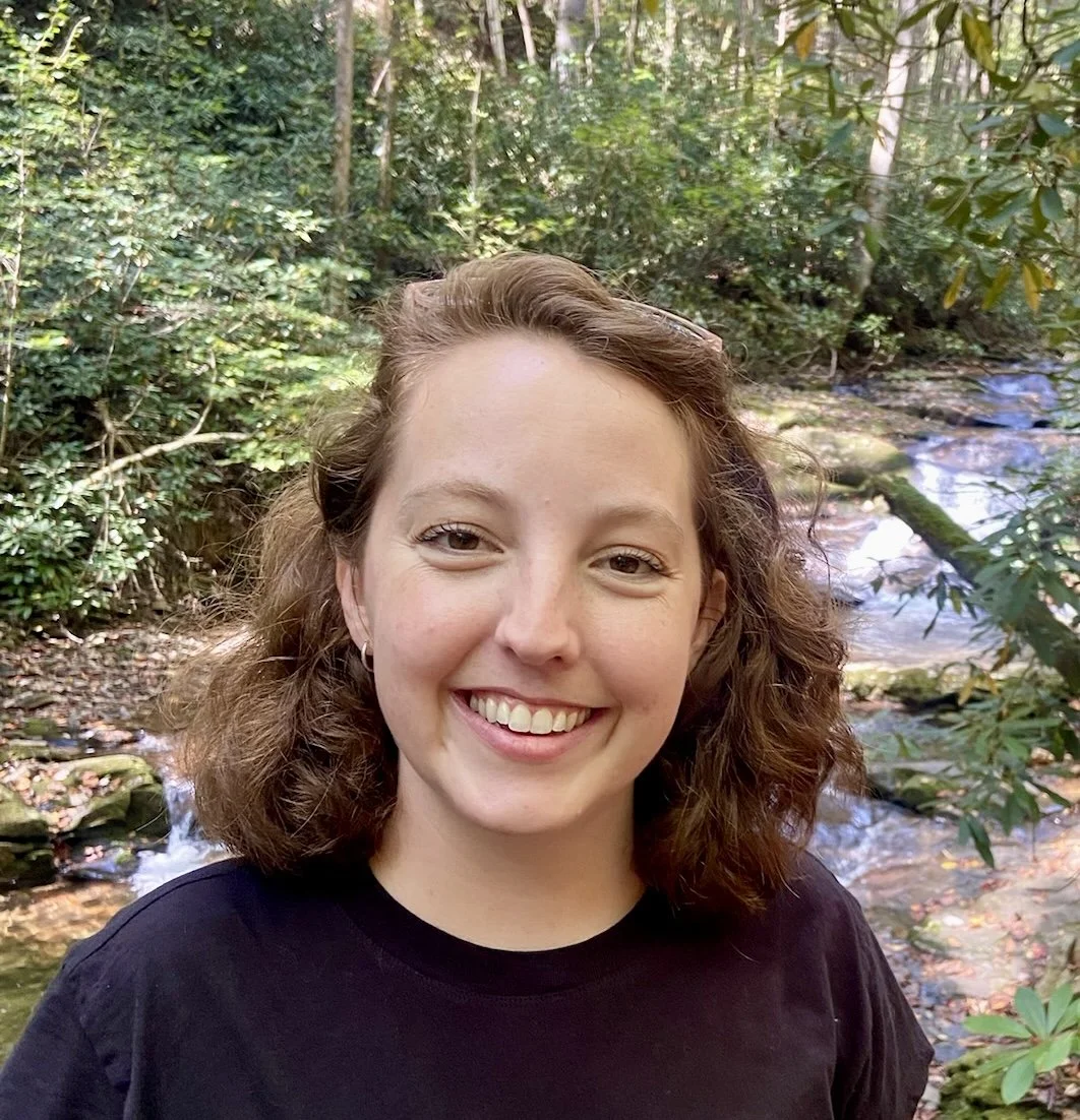 A young woman with wavy brown hair smiling outdoors near a stream in a lush green forest.