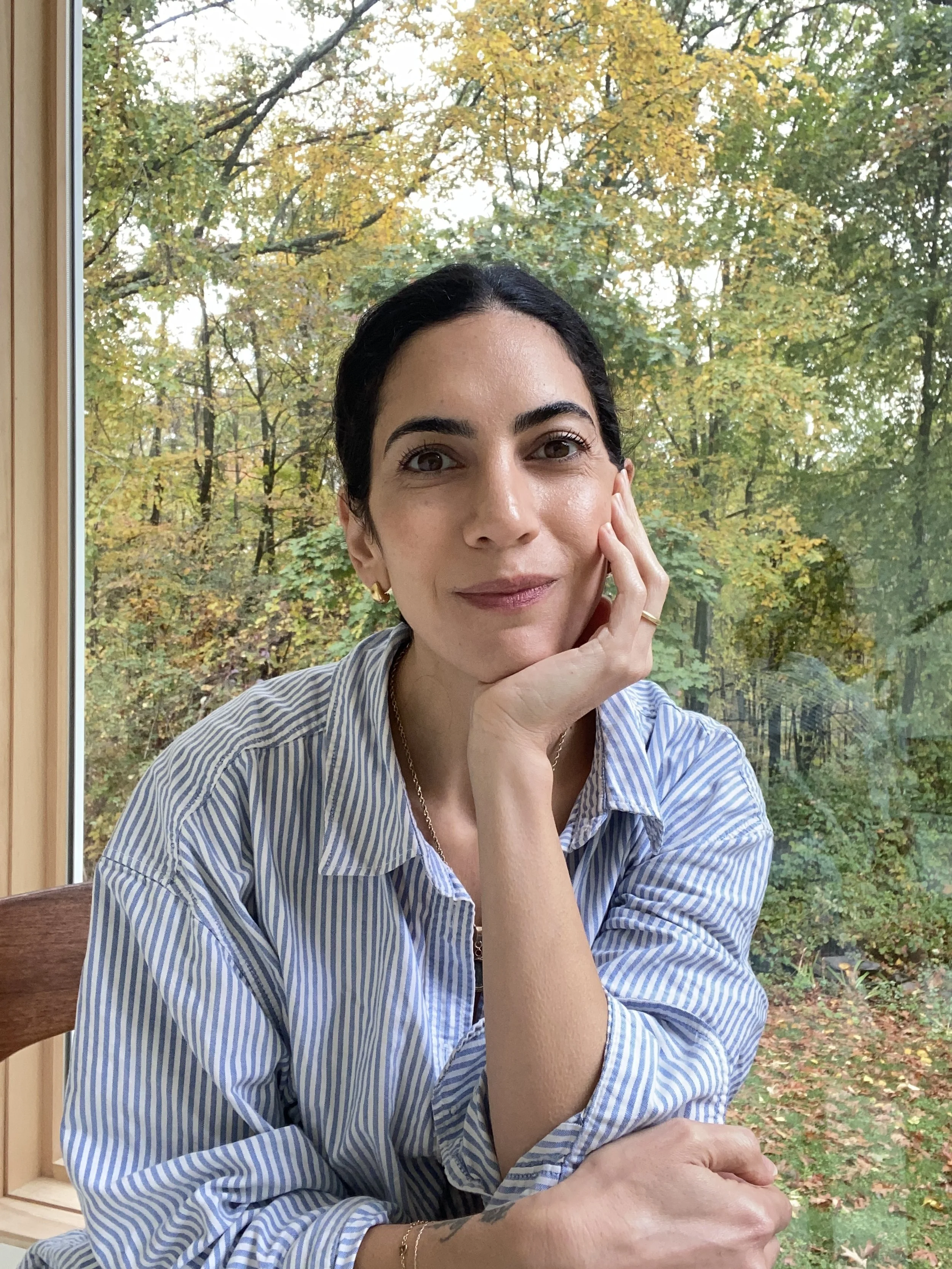A woman with dark hair and light skin sitting indoors in front of a large window showing trees with autumn leaves. She is wearing a light blue and white striped shirt and gold jewelry.