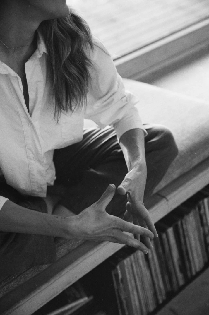 Black and white photo of a person with long hair sitting cross-legged on a bench, wearing a white shirt. The person's hand is in a mudra gesture, and the background shows a bookshelf filled with books.