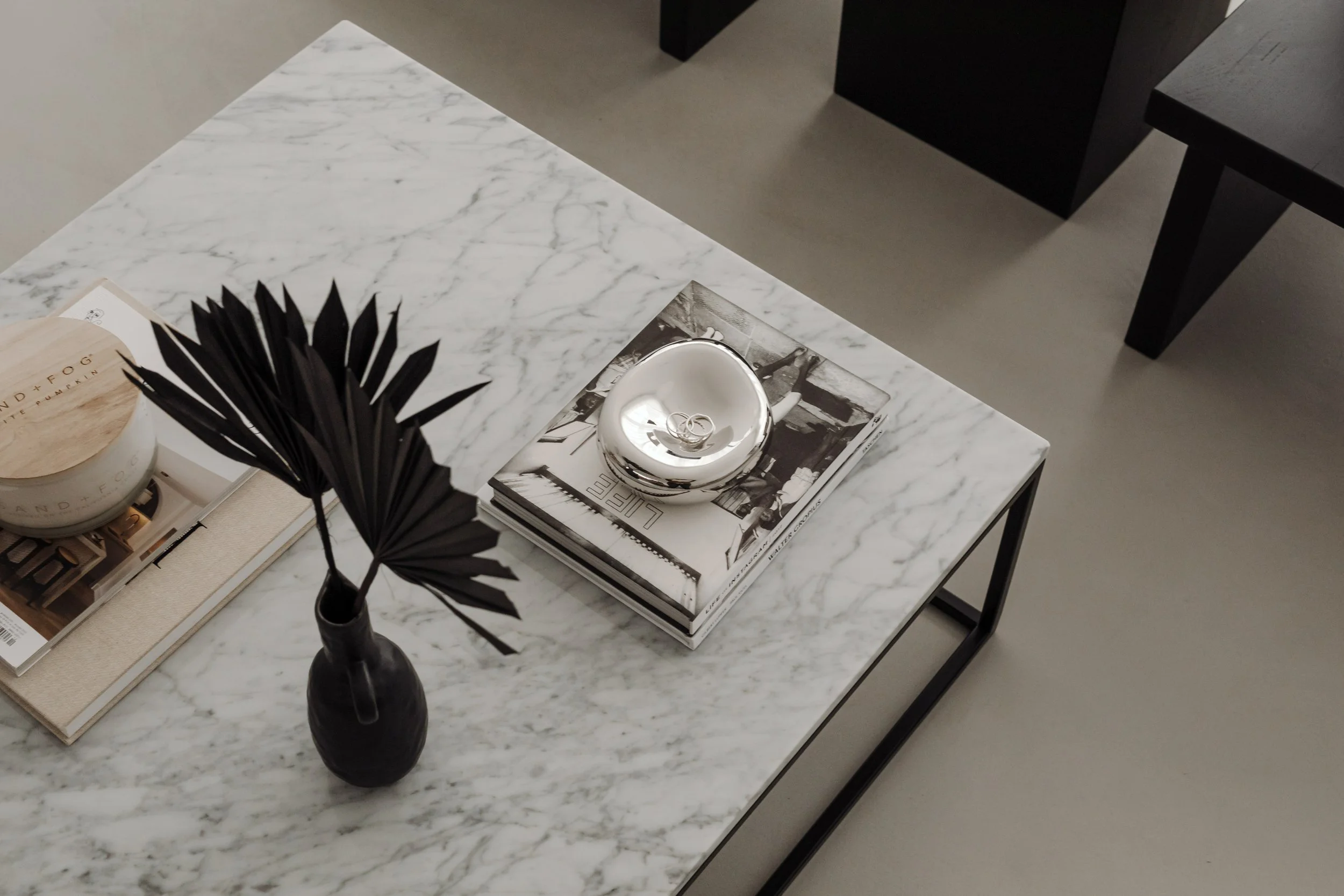 Top view of a marble coffee table with black vase holding dried black palm leaves, a black-and-white magazine with rings placed on top, and a candle on a tray, with a black chair nearby.