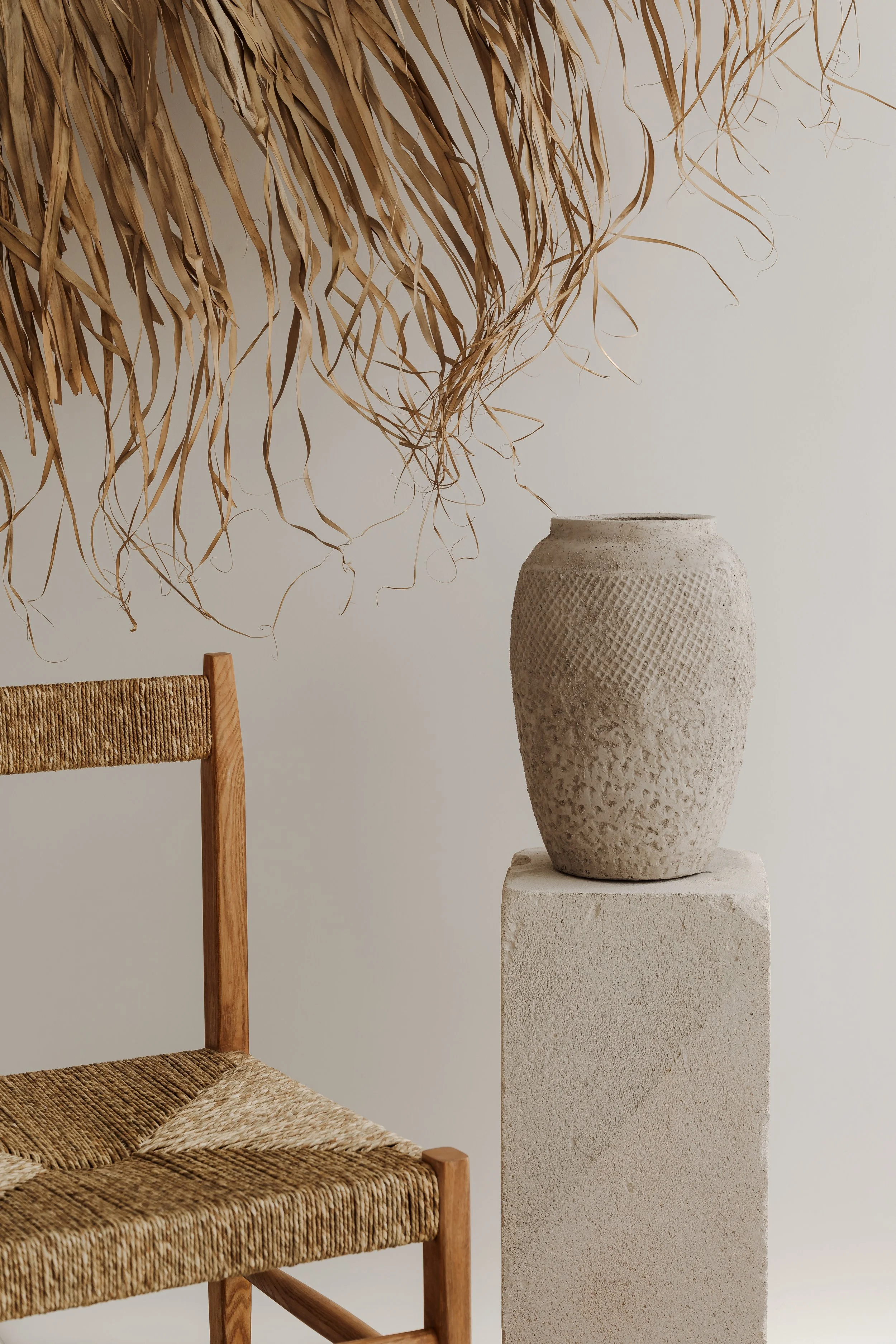 A rustic woven chair beside a white pedestal with a textured beige vase, and dried palm leaves hanging overhead against a plain white wall.
