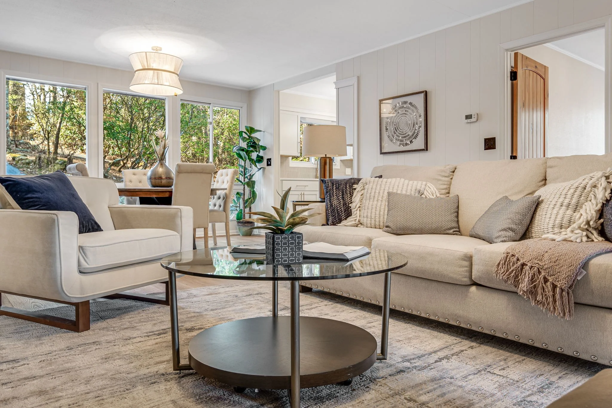 Bright living room with beige sofa and white armchair, glass coffee table, large windows with greenery outside, beige carpet, wall art, and a potted plant.