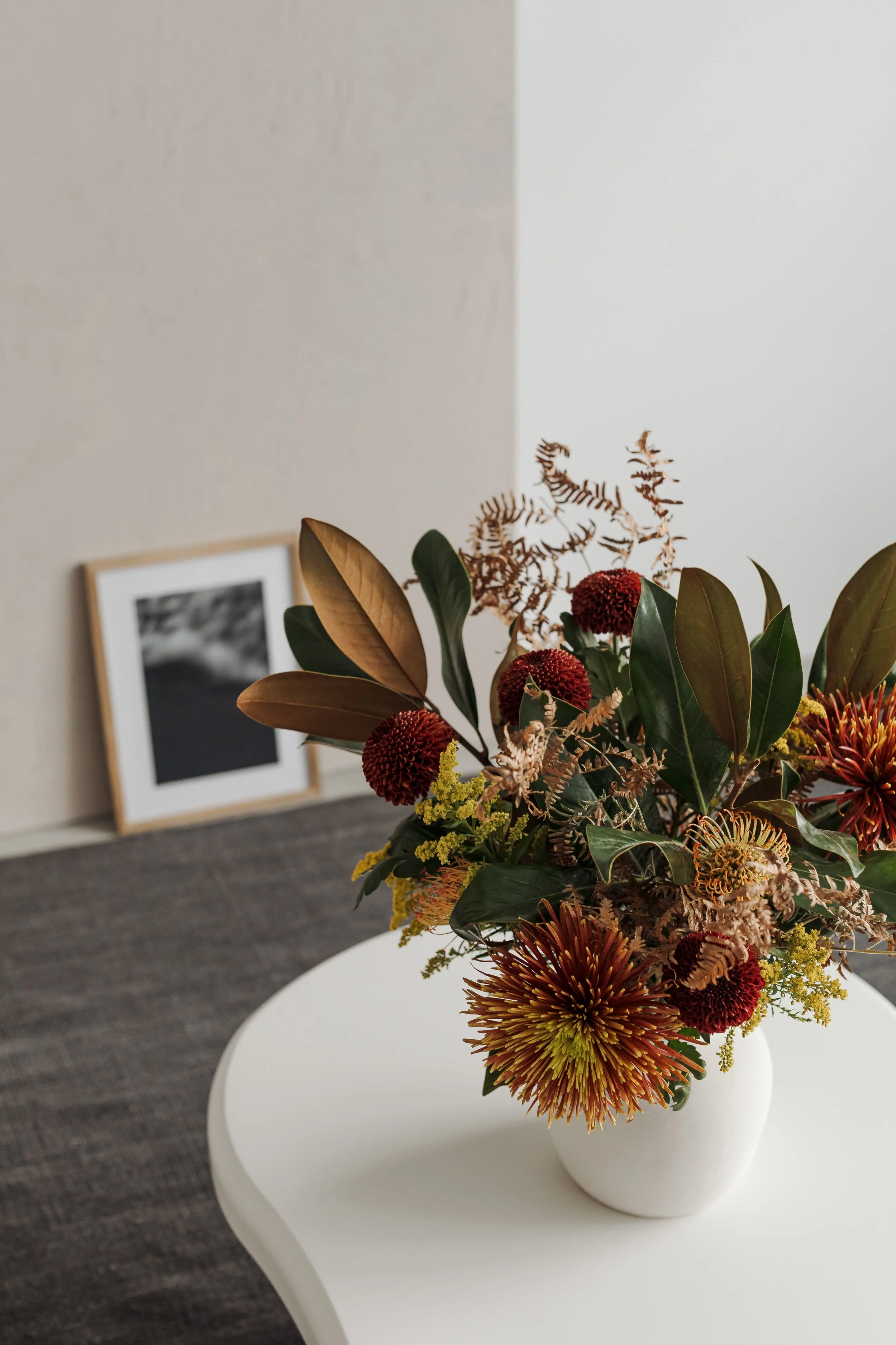 A floral arrangement in a white vase on a white table, with a black and white framed photograph in the background.