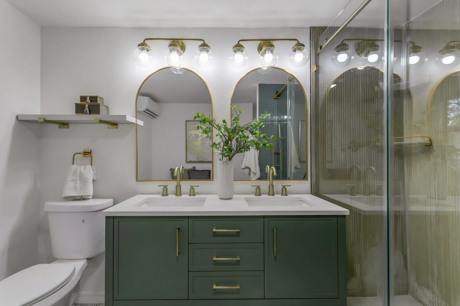 Modern bathroom with a double sink vanity, gold fixtures, two oval mirrors, a potted plant, and a glass shower enclosure.