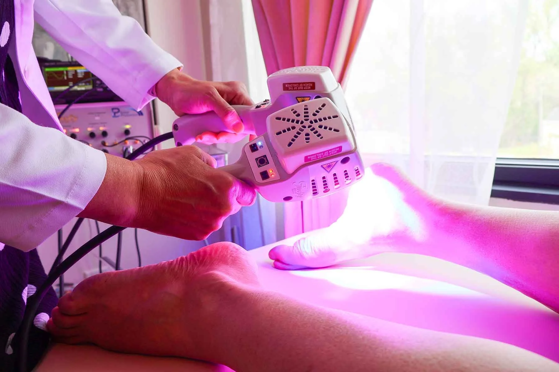 A medical professional uses a laser therapy device on a patient's arm in a well-lit room with pink curtains and a window in the background.
