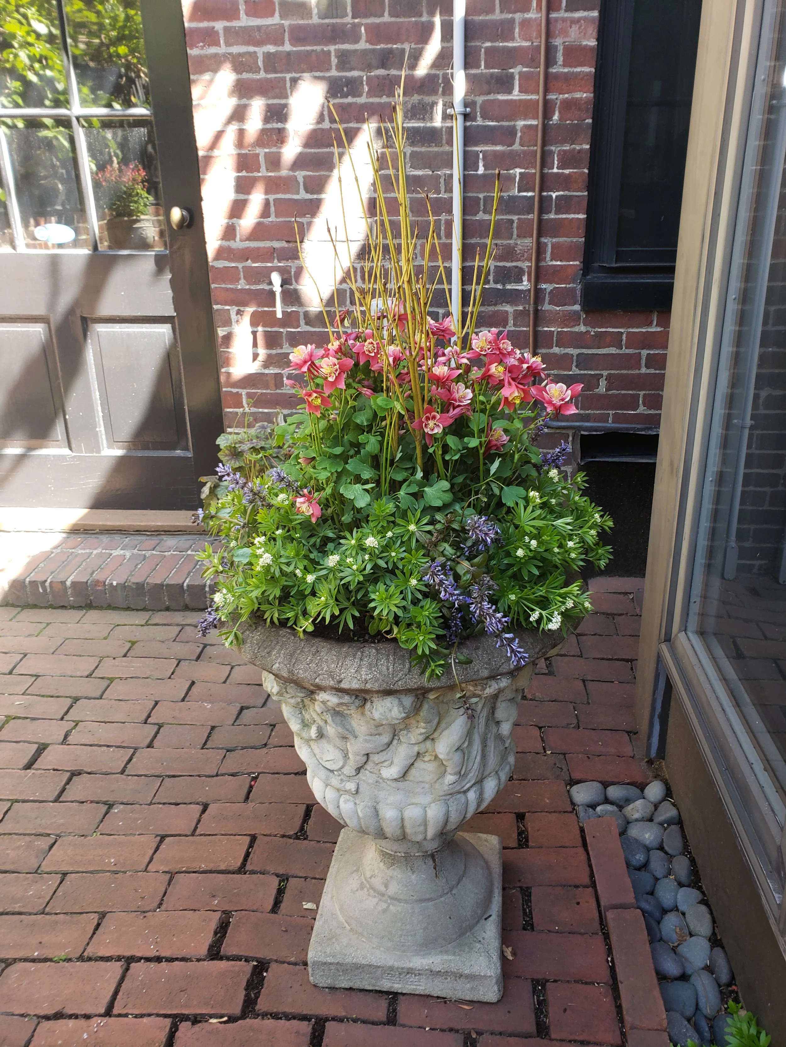 Decorative stone planter filled with pink, purple, and white flowering plants, placed on a brick patio outside a brick house.