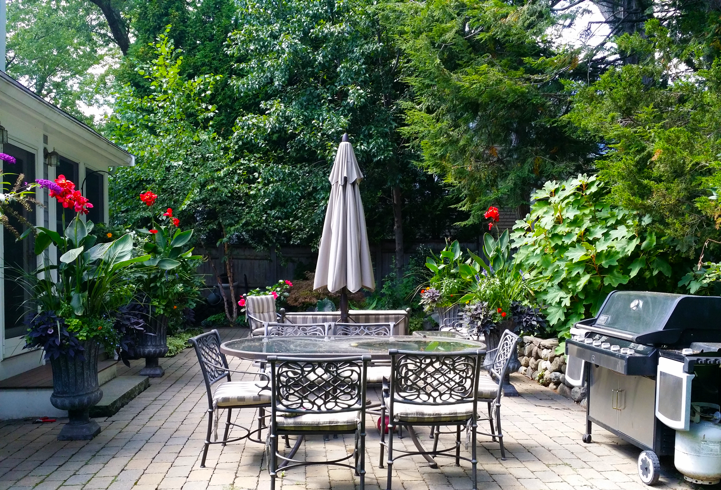 A backyard patio with a glass table and six metal chairs around it, a large patio umbrella in the center, a propane grill on the right, and lush green plants and trees in the background.
