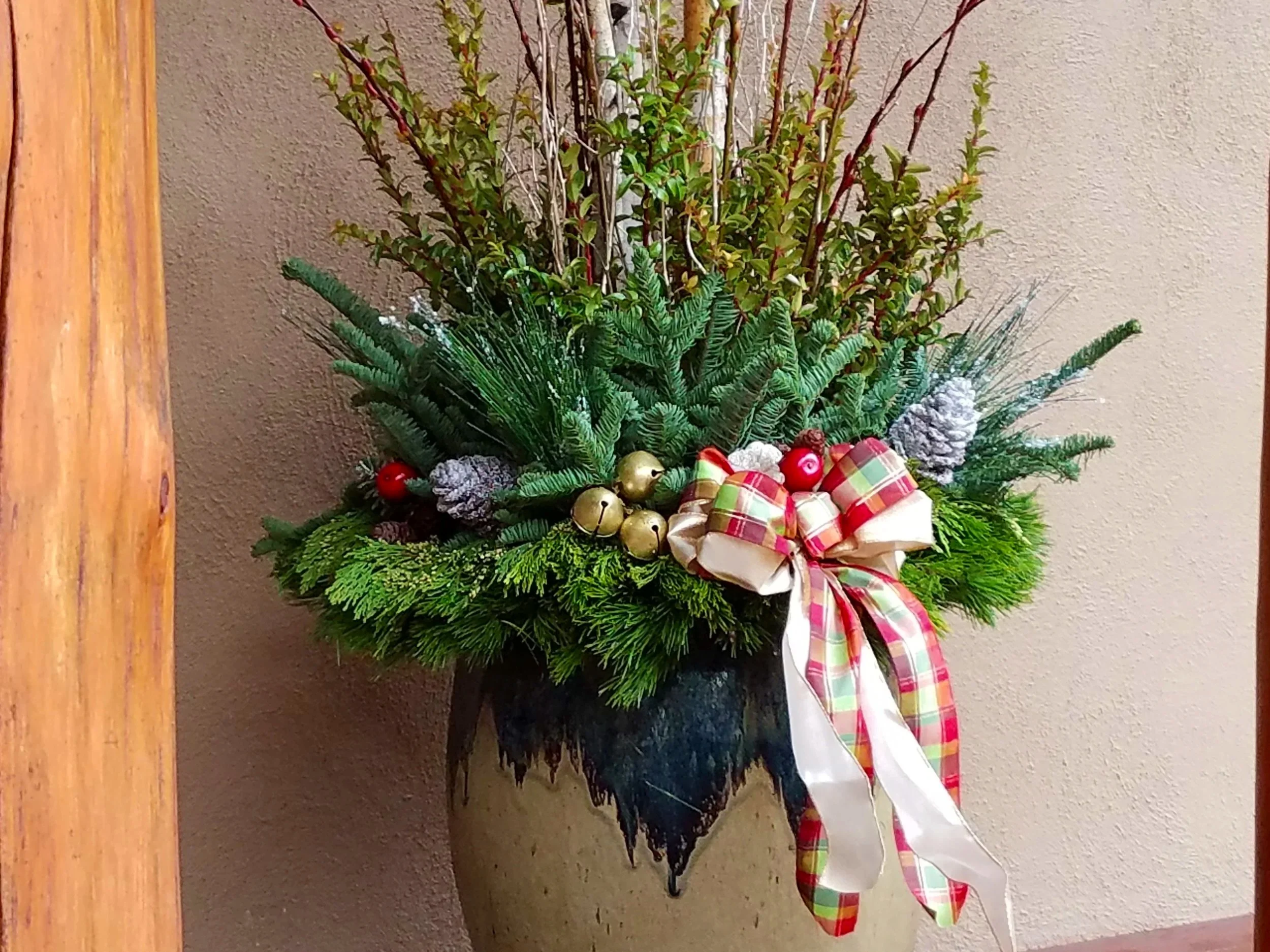 A decorative Christmas arrangement with pine branches, red berries, pinecones, gold ornaments, and a plaid ribbon, placed in a large ceramic pot against a beige wall.