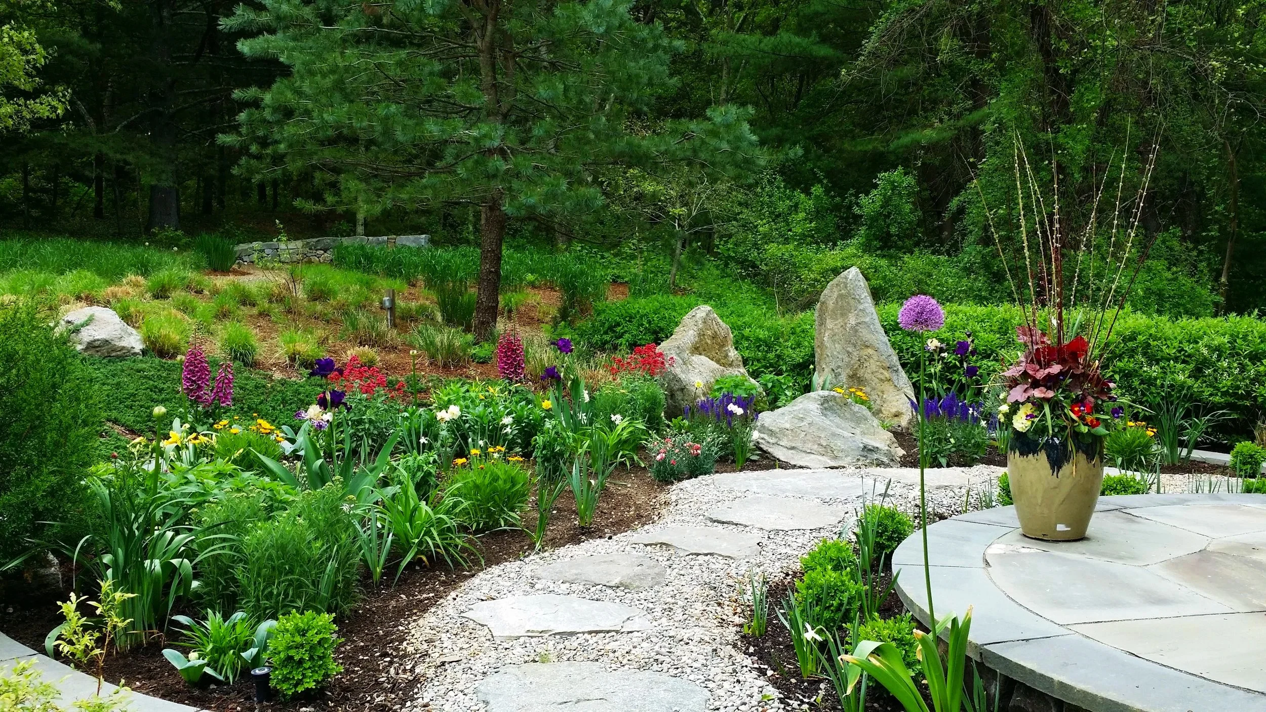 A well-maintained garden with a stone pathway, colorful flowers including purple, yellow, and red blooms, a large potted plant with purple flowers on a circular stone platform, and a background of lush green trees and shrubs.