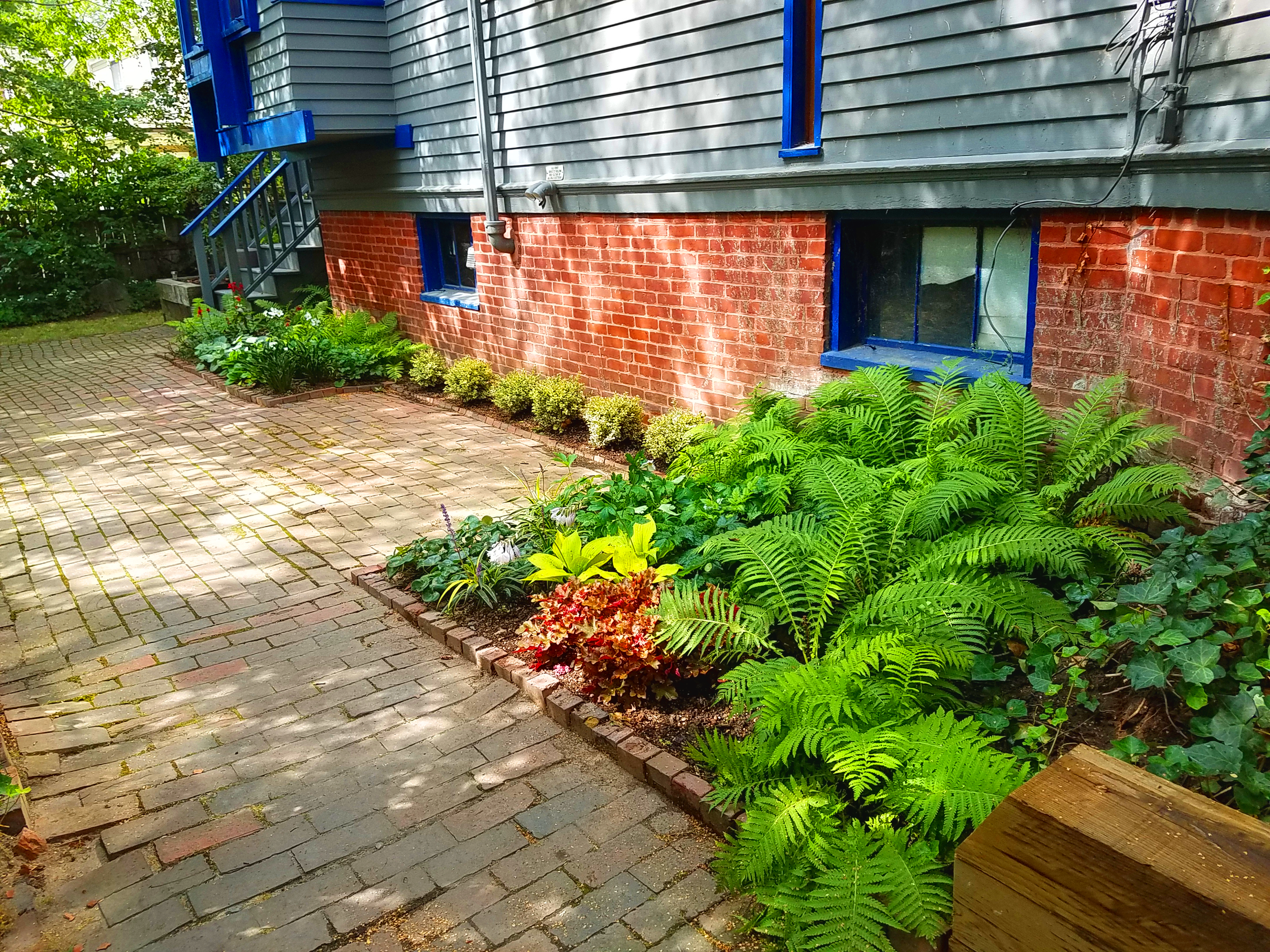 A brick house with gray siding and blue window frames, with a small garden and brick-paved pathway in the front yard. There are various green plants and flowers along the garden bed.