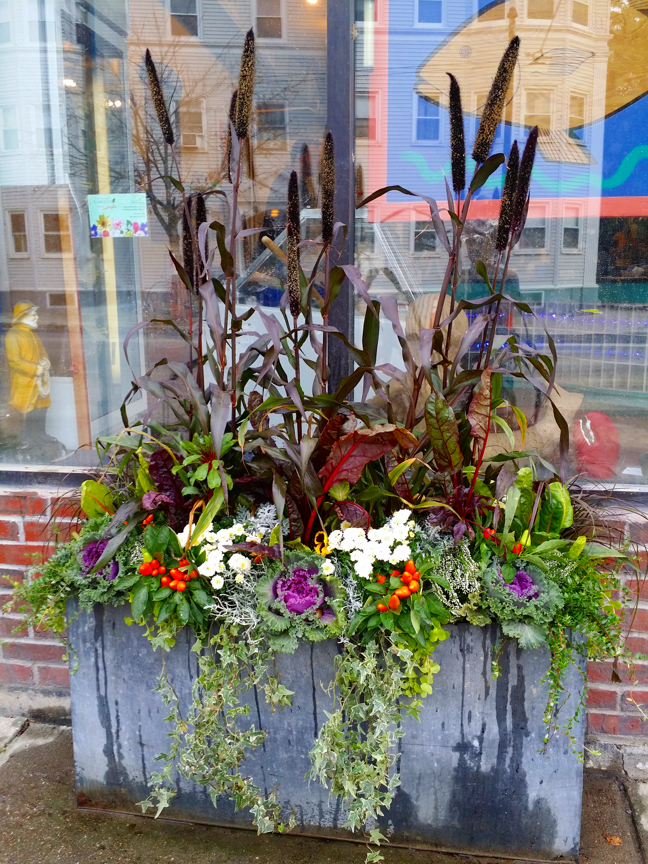 Decorative fall display of various plants and flowers in a large gray container outside a window, with a city street scene reflected in the glass.