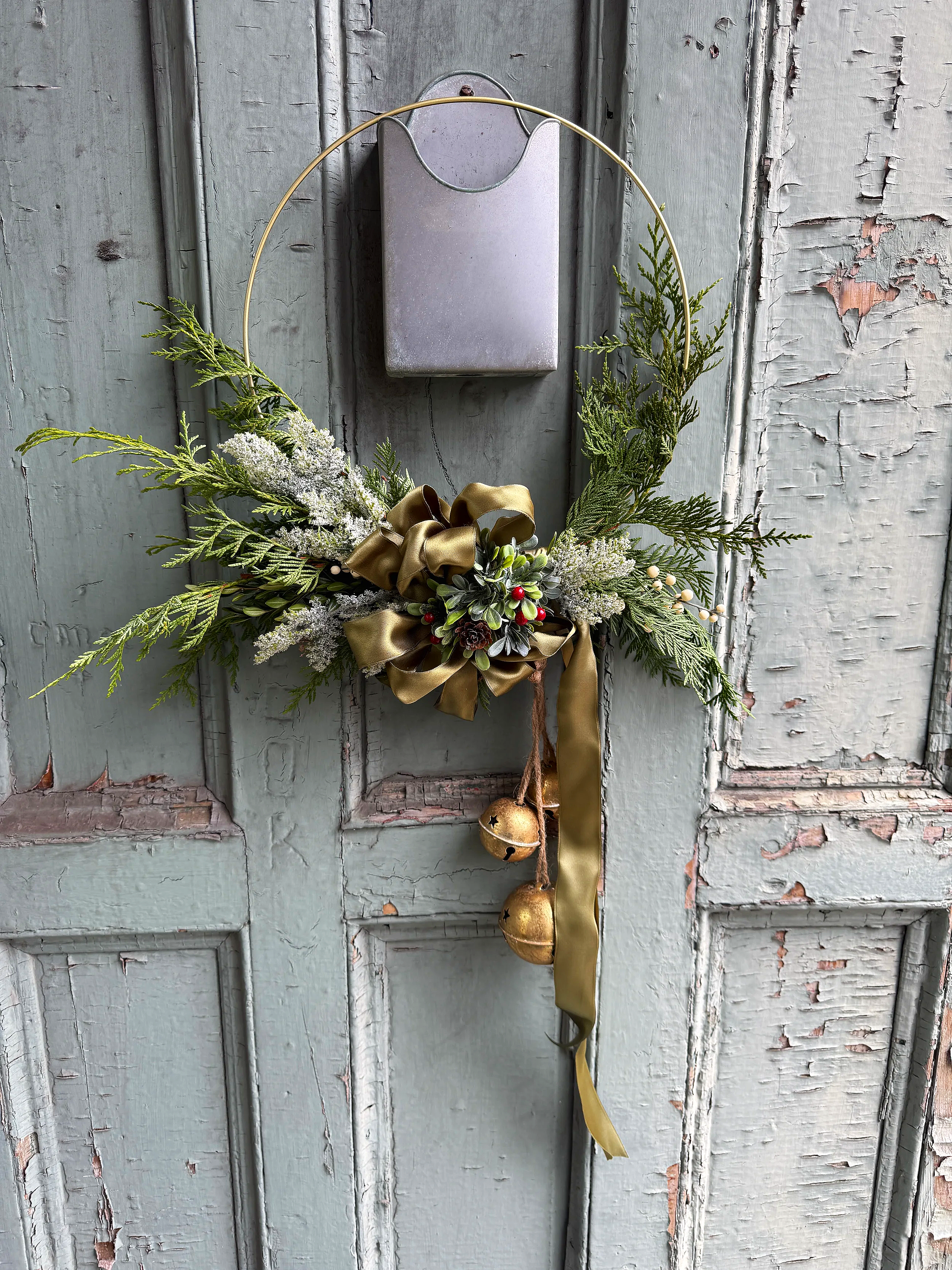 Decorative holiday wreath with greenery, gold ribbon, small ornaments, and a bow, hanging on a weathered light green door.