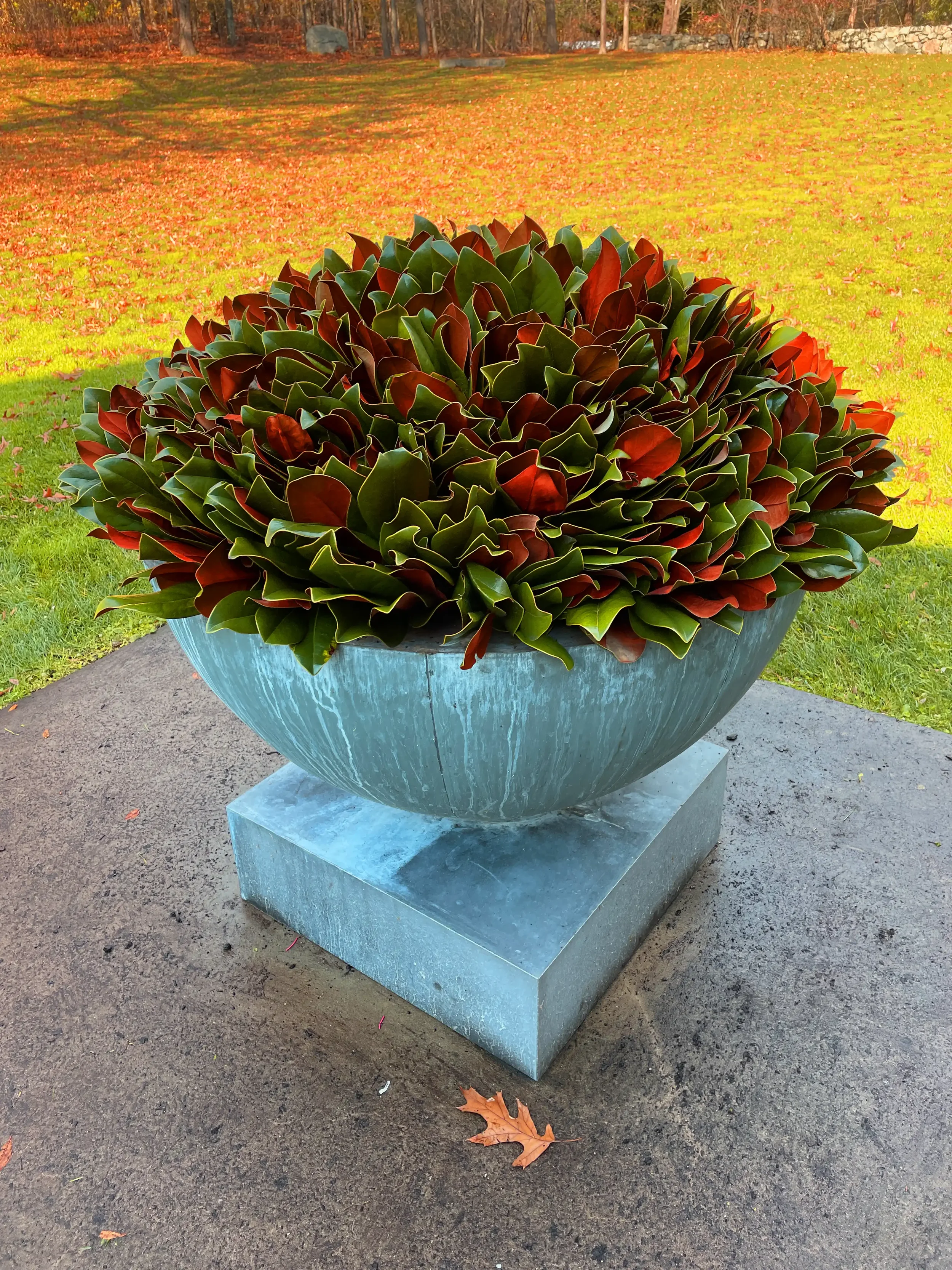 A large round planter filled with green and red-leaved plants is placed on a square stone base on a concrete surface outdoors with an autumn-colored lawn and trees in the background.