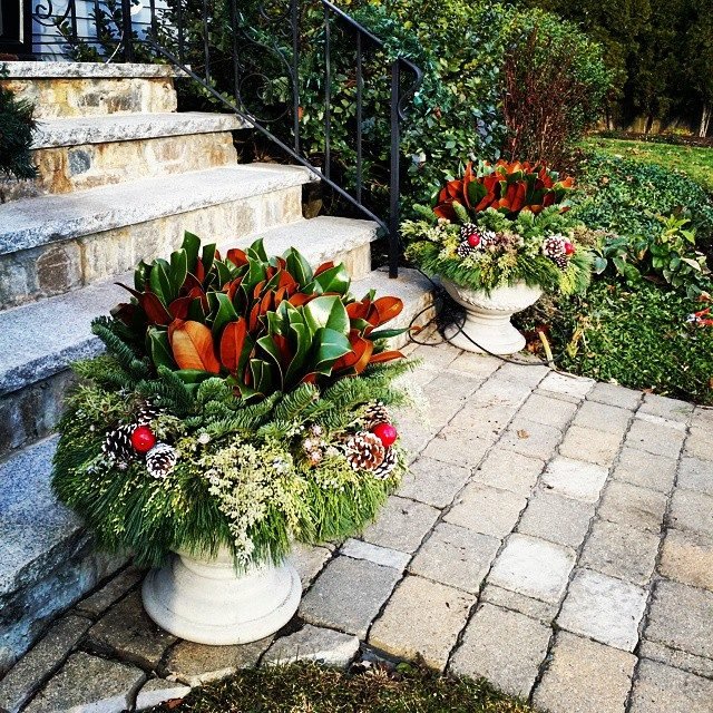 Two decorative planters with holiday floral arrangements placed on a stone patio near a staircase with stone steps and a black metal railing, surrounded by greenery.