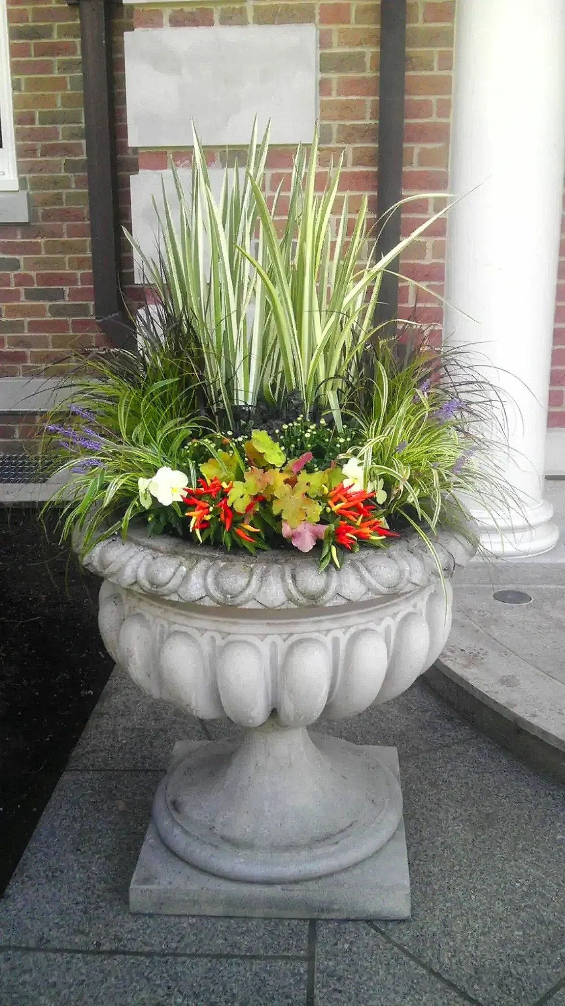 A large decorative stone planter filled with various green plants and colorful flowers, situated outside a building with brick walls and columns.