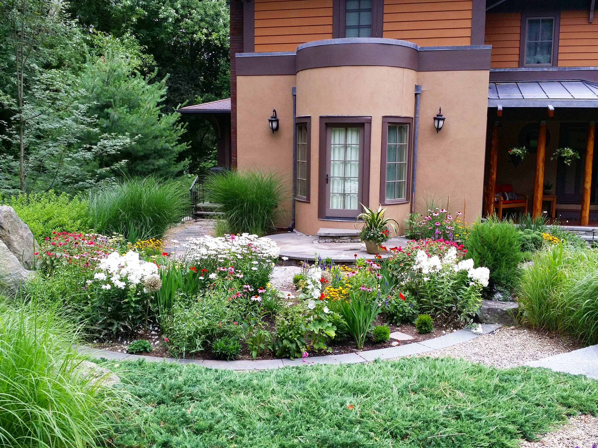 A house with a landscaped garden featuring various blooming flowers, green shrubs, and trees, with a pathway leading to the front door.