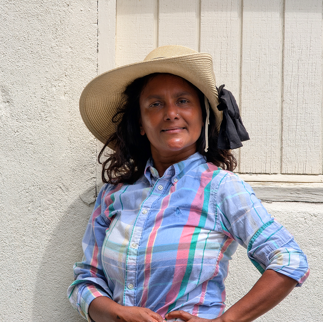 A woman wearing a wide-brimmed straw hat with a black ribbon, a blue plaid button-up shirt, standing against a textured white wall with a window behind her.