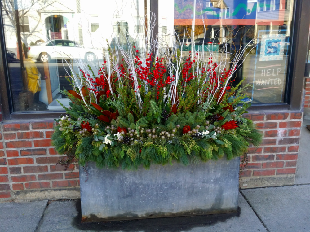 A large rectangular concrete planter filled with holiday-themed greenery and decorations, including red berries, white twigs, and assorted green foliage, placed outside a brick storefront with glass windows.