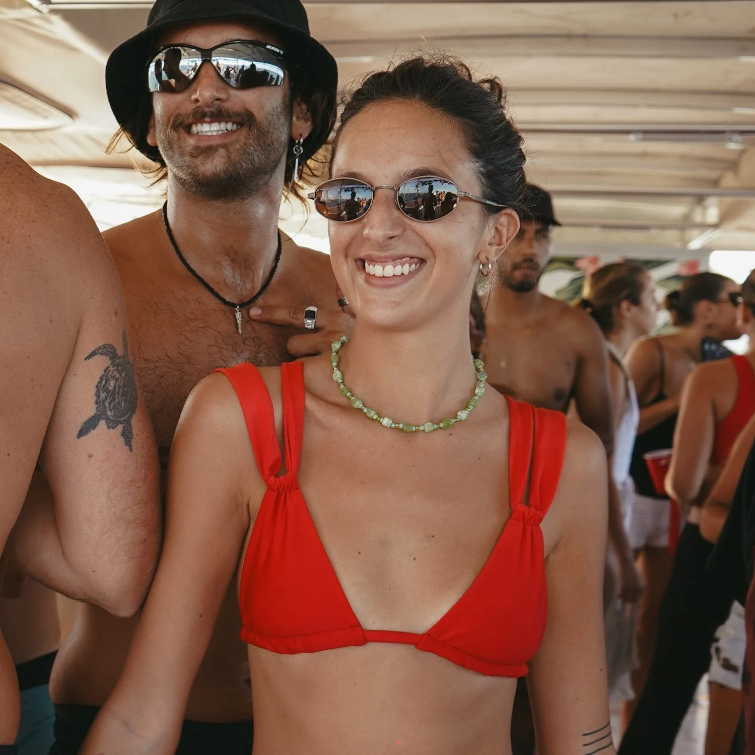 Smiling group of young adults at a party or gathering, with a focus on a woman in a red top and sunglasses, and a man with sunglasses and a beard, wearing a hat, in a social setting with others in the background.