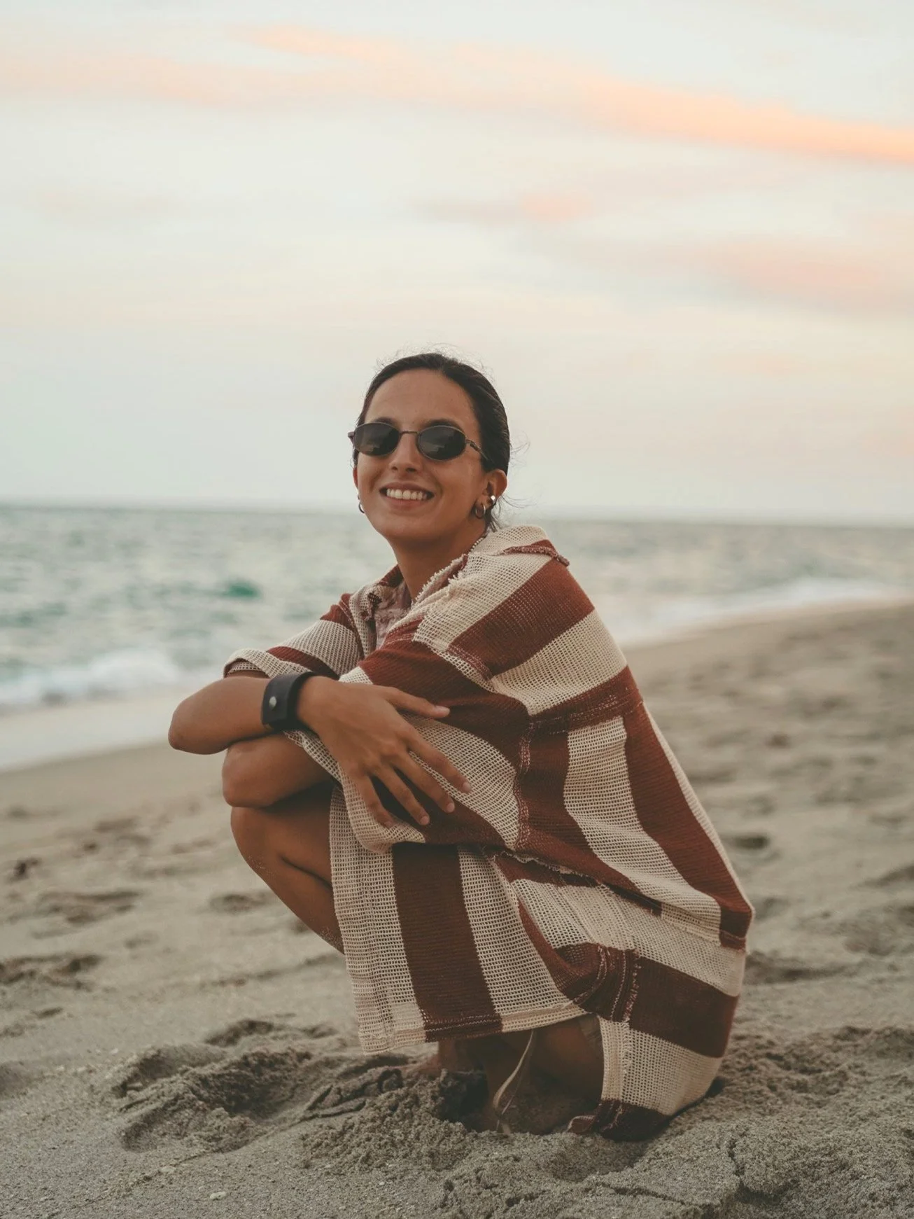 A woman sitting on the sandy beach near the ocean, smiling, wearing sunglasses, a striped shawl, and a watch, during sunset with pink and orange clouds.