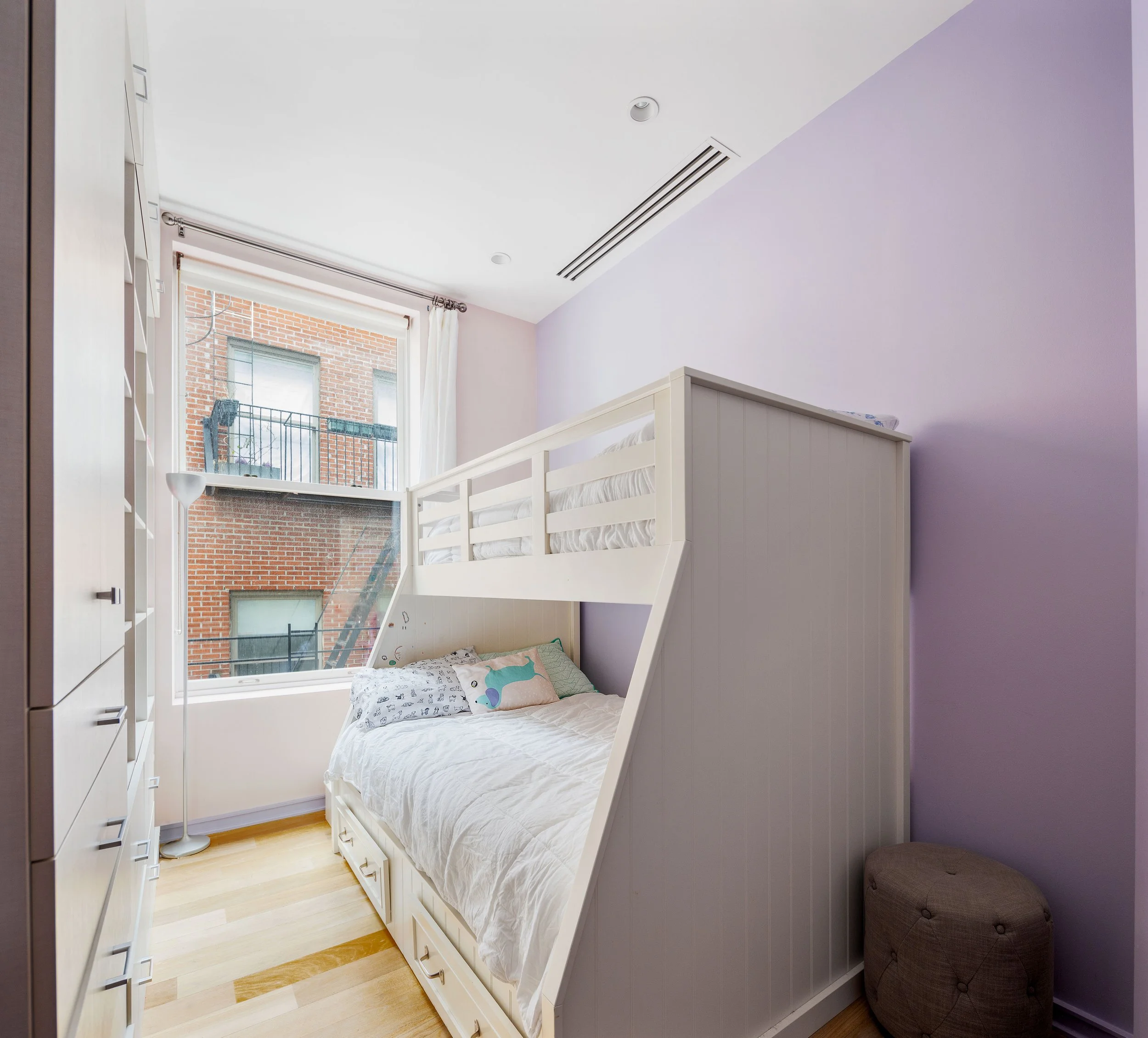 A small bedroom with a white bunk bed with a lower bed and an upper bed, positioned near a window with white curtains. The room has a lavender-colored wall, light wood flooring, a brown cushioned ottoman, and a floor lamp near the window.