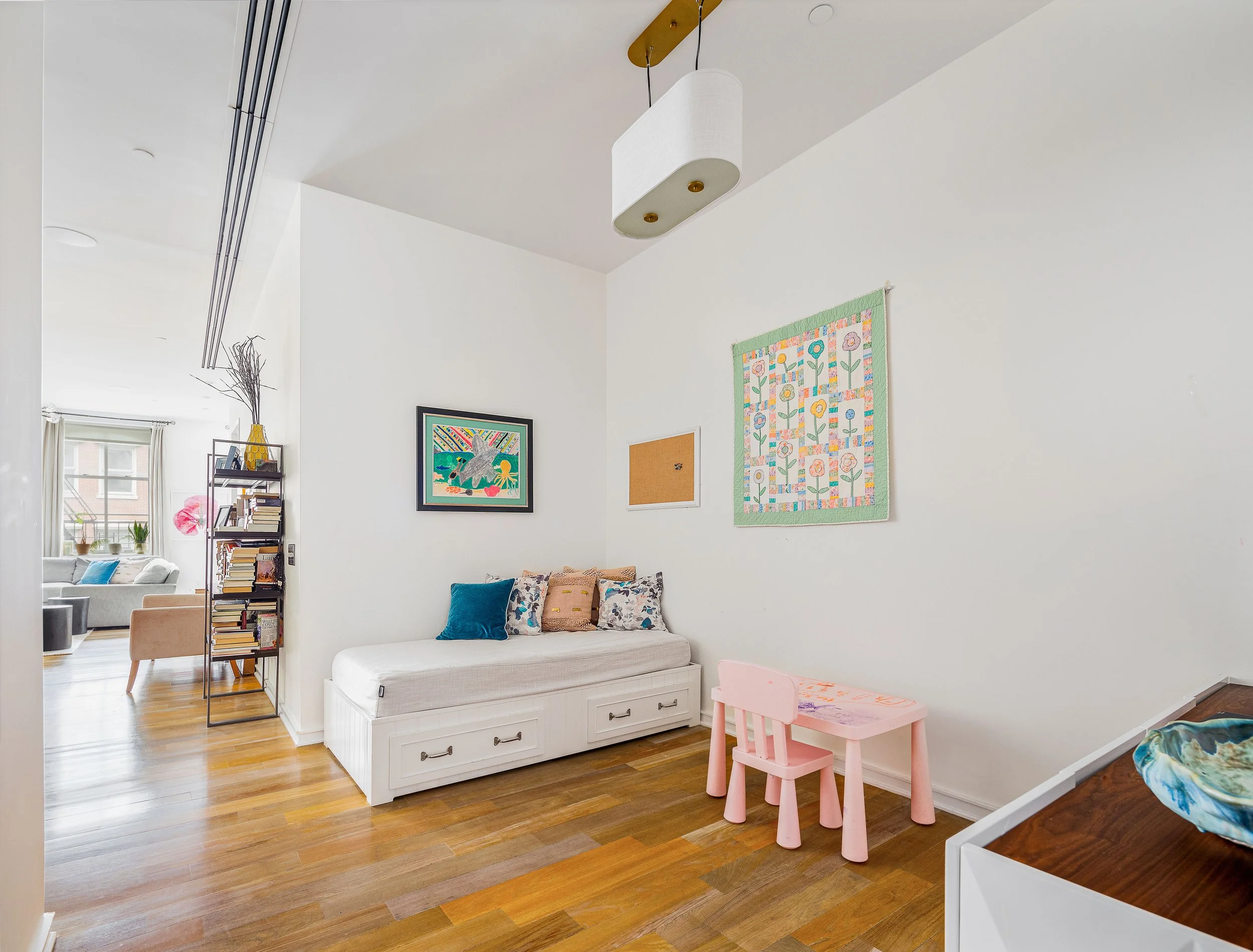 Living room with white walls, wooden flooring, a white bench with storage drawers and colorful cushions, a small pink children's table and chair, children's artwork on the wall, and a bookshelf with books, leading to a sitting area with a gray sofa a