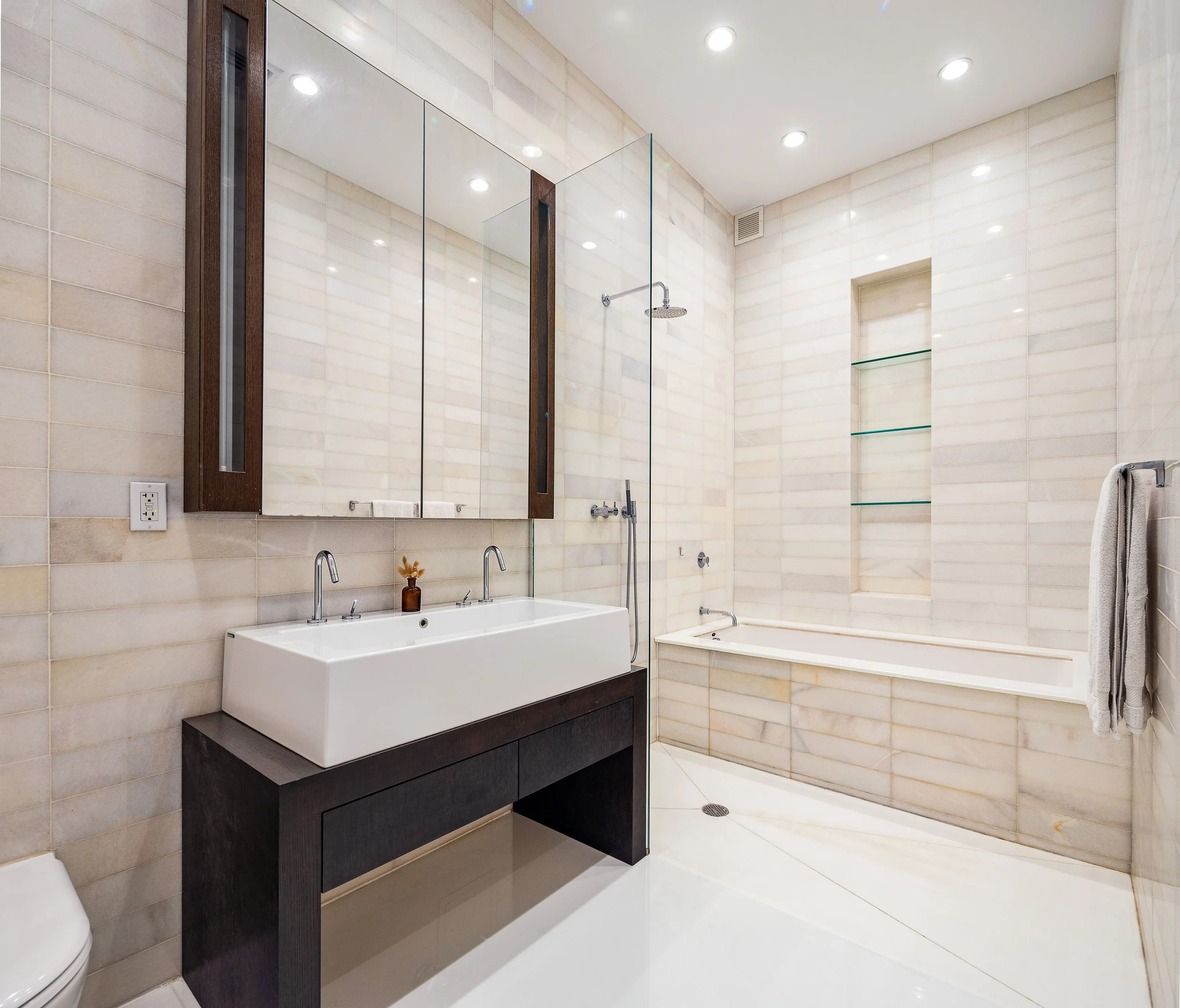 A modern bathroom with a double sink vanity, large mirror, glass-enclosed shower, built-in bathtub, and a neutral tile design.