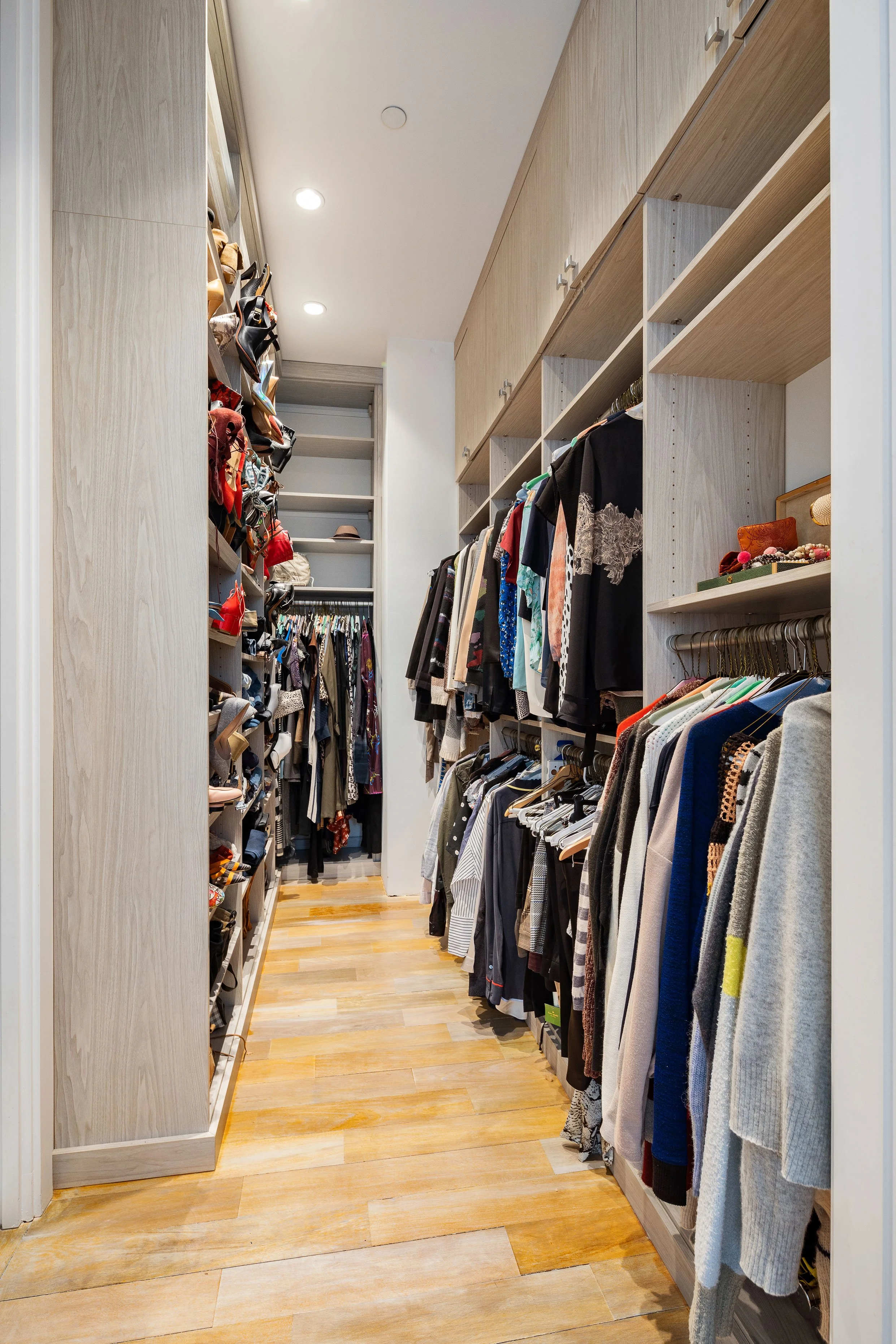 A walk-in closet with wooden floor and built-in shelves. Clothing and accessories are organized on hanging rods and shelves on either side, with the space narrowing towards the back.