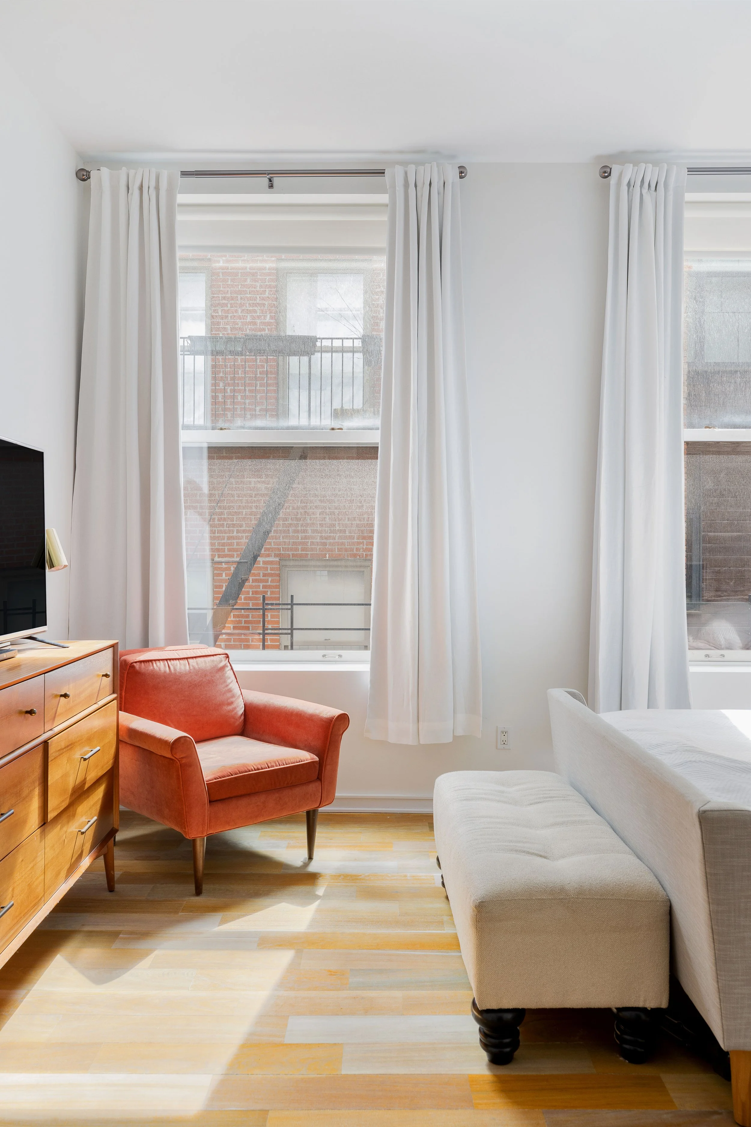 Bright living room with two large windows with white curtains, a red armchair, a beige chaise lounge, and a wooden dresser with a small TV.