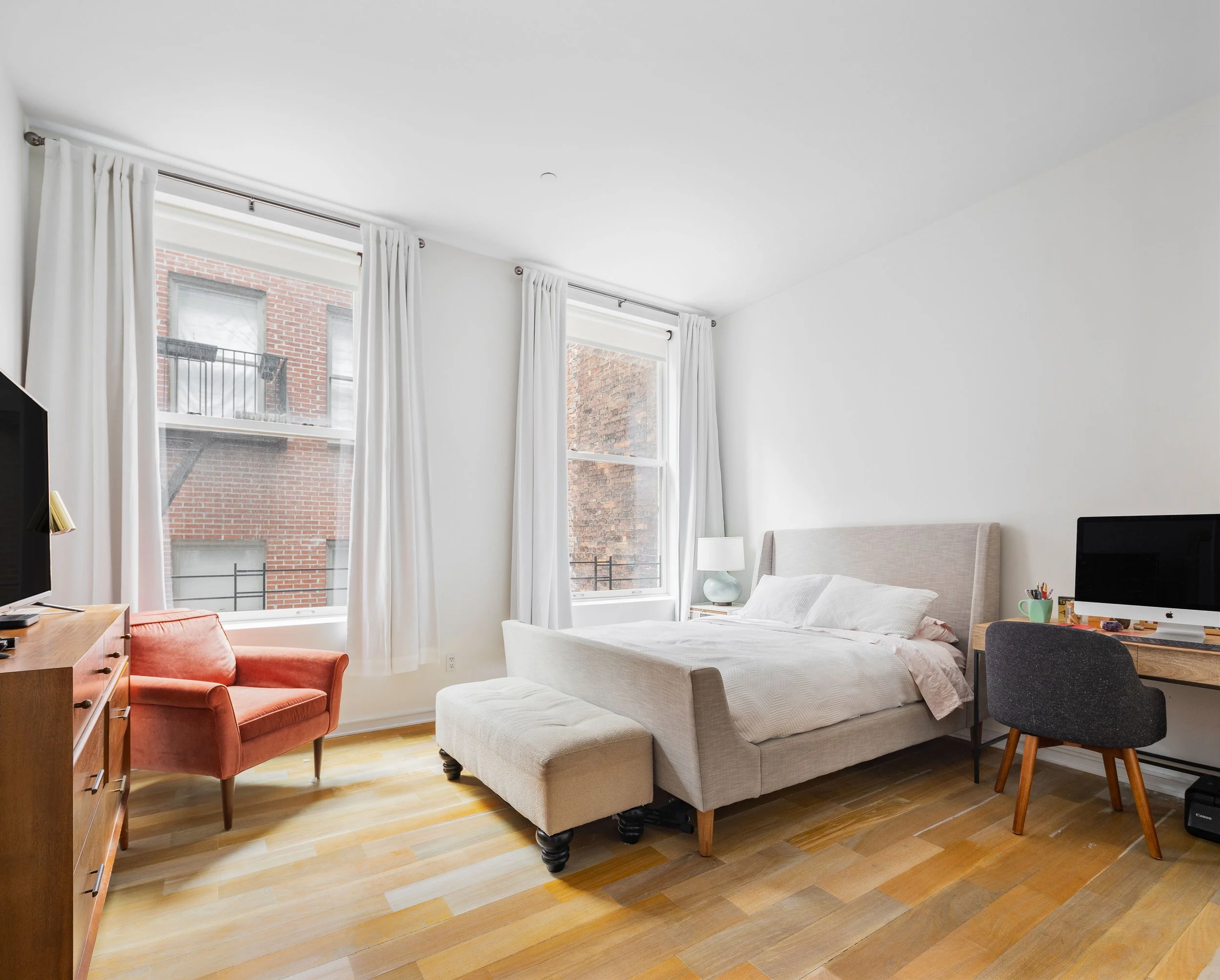 Bright bedroom with large windows, white walls, beige bed, nightstand with lamp, orange armchair, desk with computer, and wooden floors.