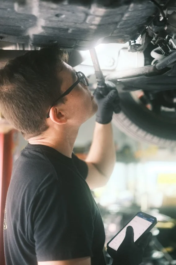 A technician is using a flashlight to observe the underside of a vehicle while performing an inspection